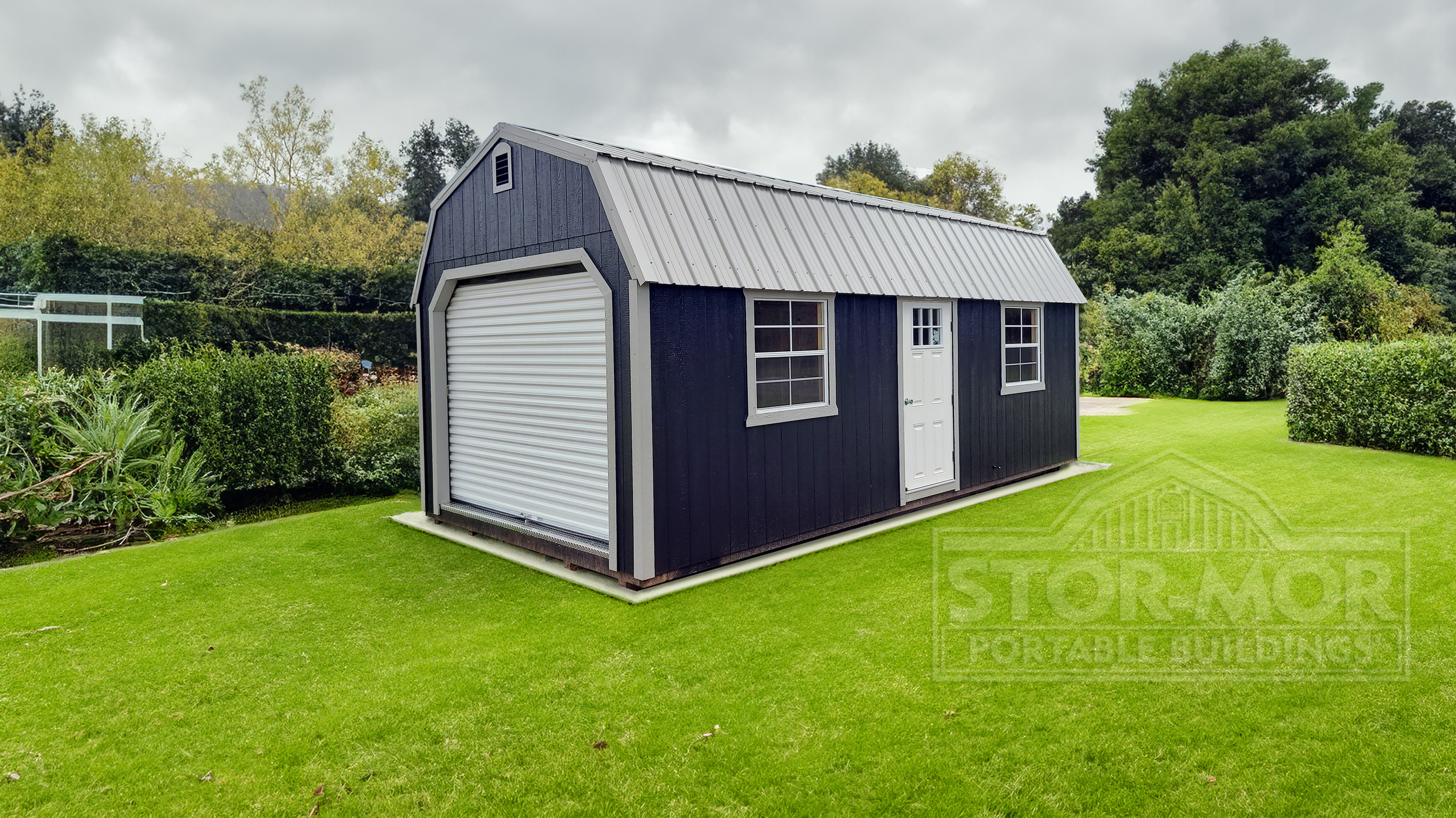 A dark blue metal shed with a white roll-up garage door, a small white door with a window, and two large windows on a well-maintained green lawn, surrounded by trees and bushes under an overcast sky.