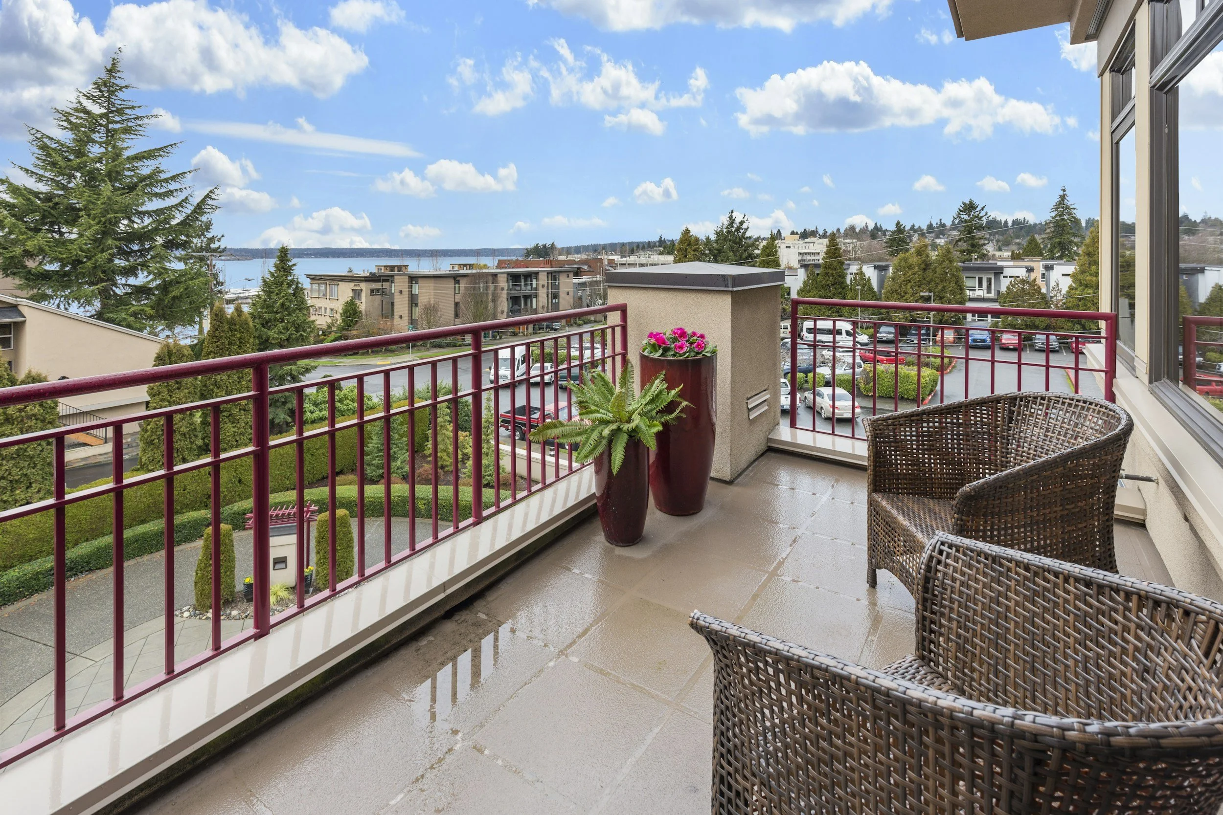 Balcony with wicker chairs and potted plants, overlooking a residential area with trees, parked cars, and water in the background, under a partly cloudy sky.