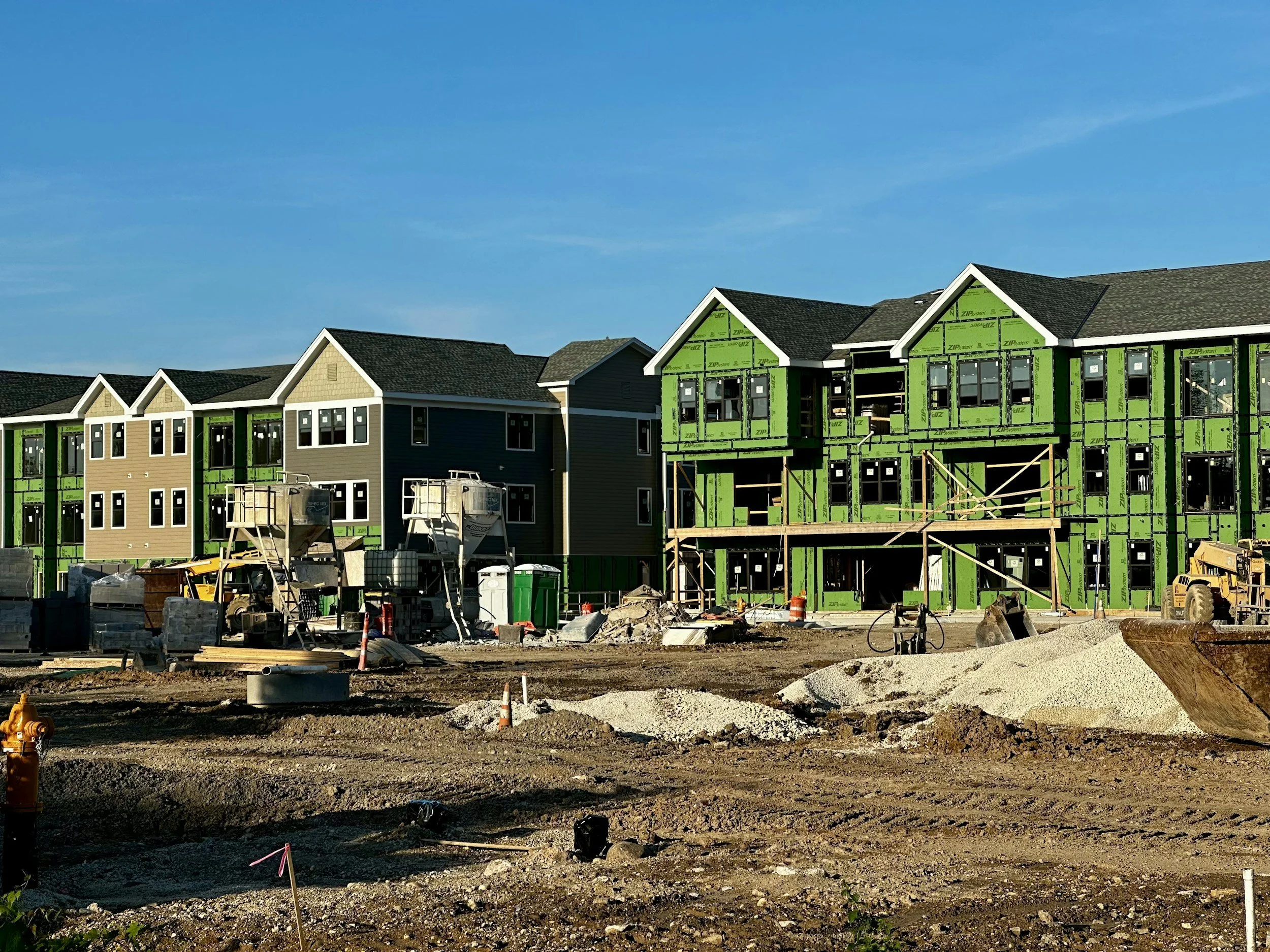 Under-construction houses with partially installed exterior walls, scaffolding, construction equipment, and a dirt lot under a clear blue sky.