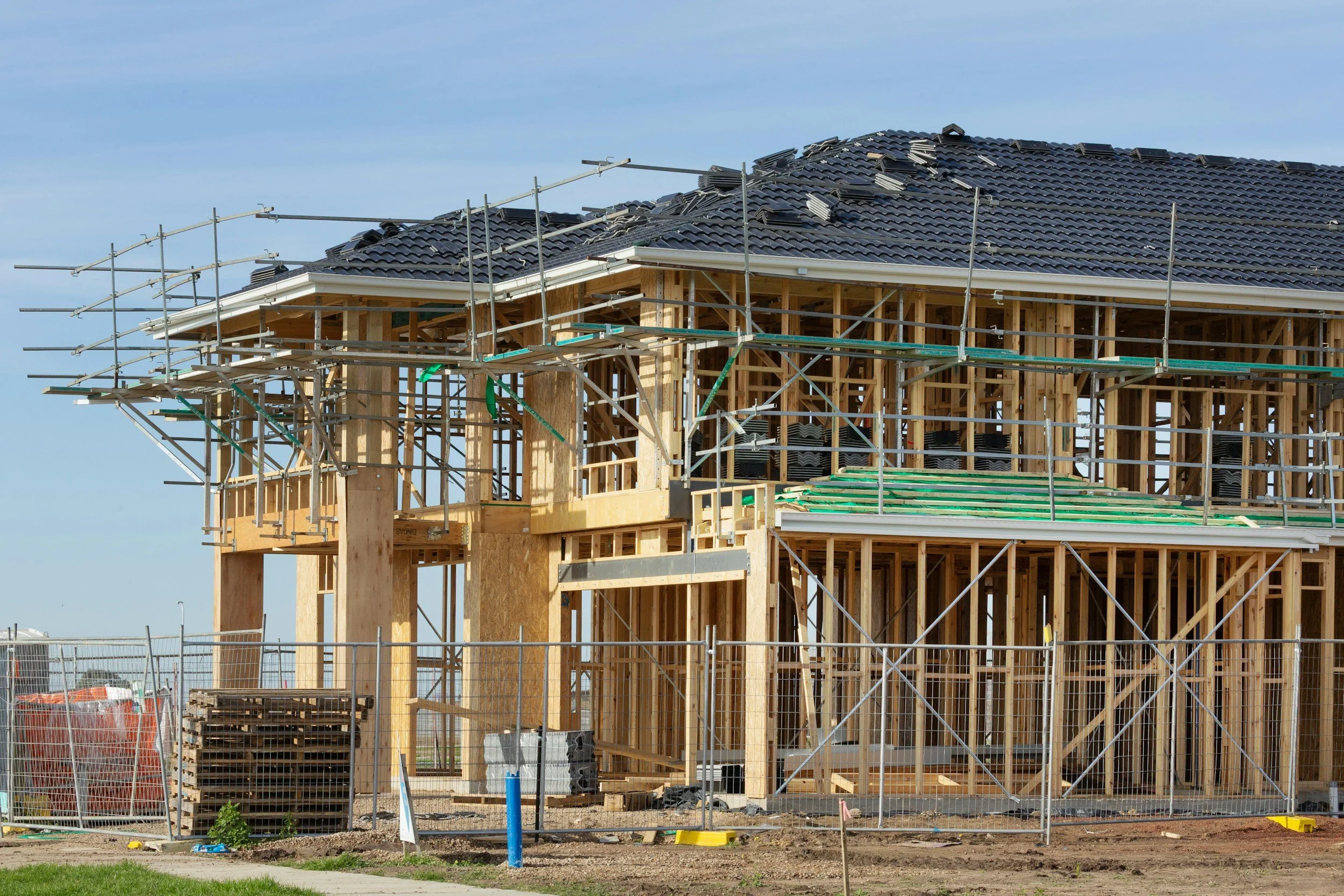 Construction site with a wooden frame of a building, scaffolding around it, and a partially completed roof with black tiles, under a clear blue sky.