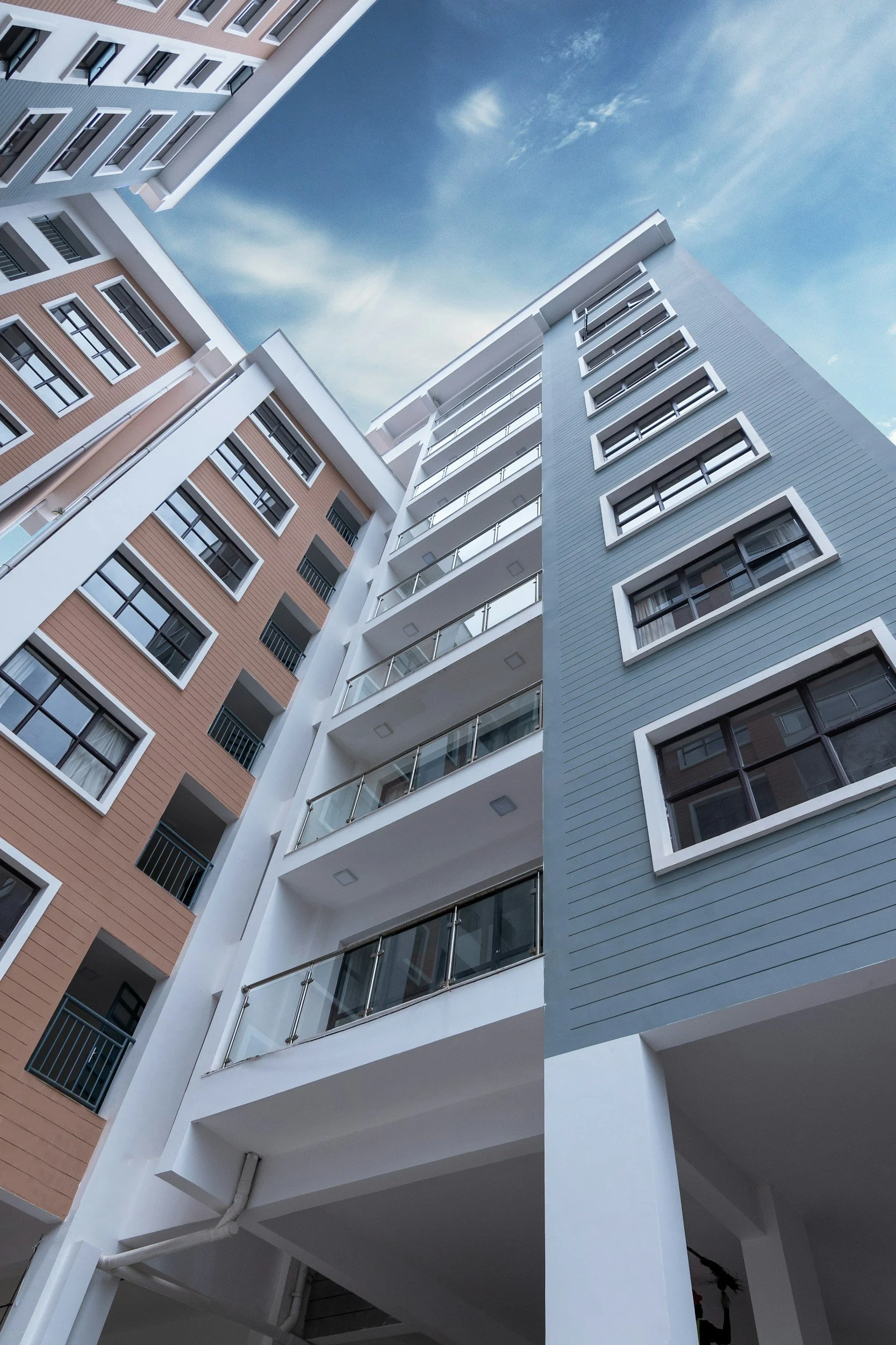 A modern multi-story apartment building with pastel pink and blue exteriors, white balcony railings, and large windows. The photo is taken from below, with a blue sky and some clouds overhead.