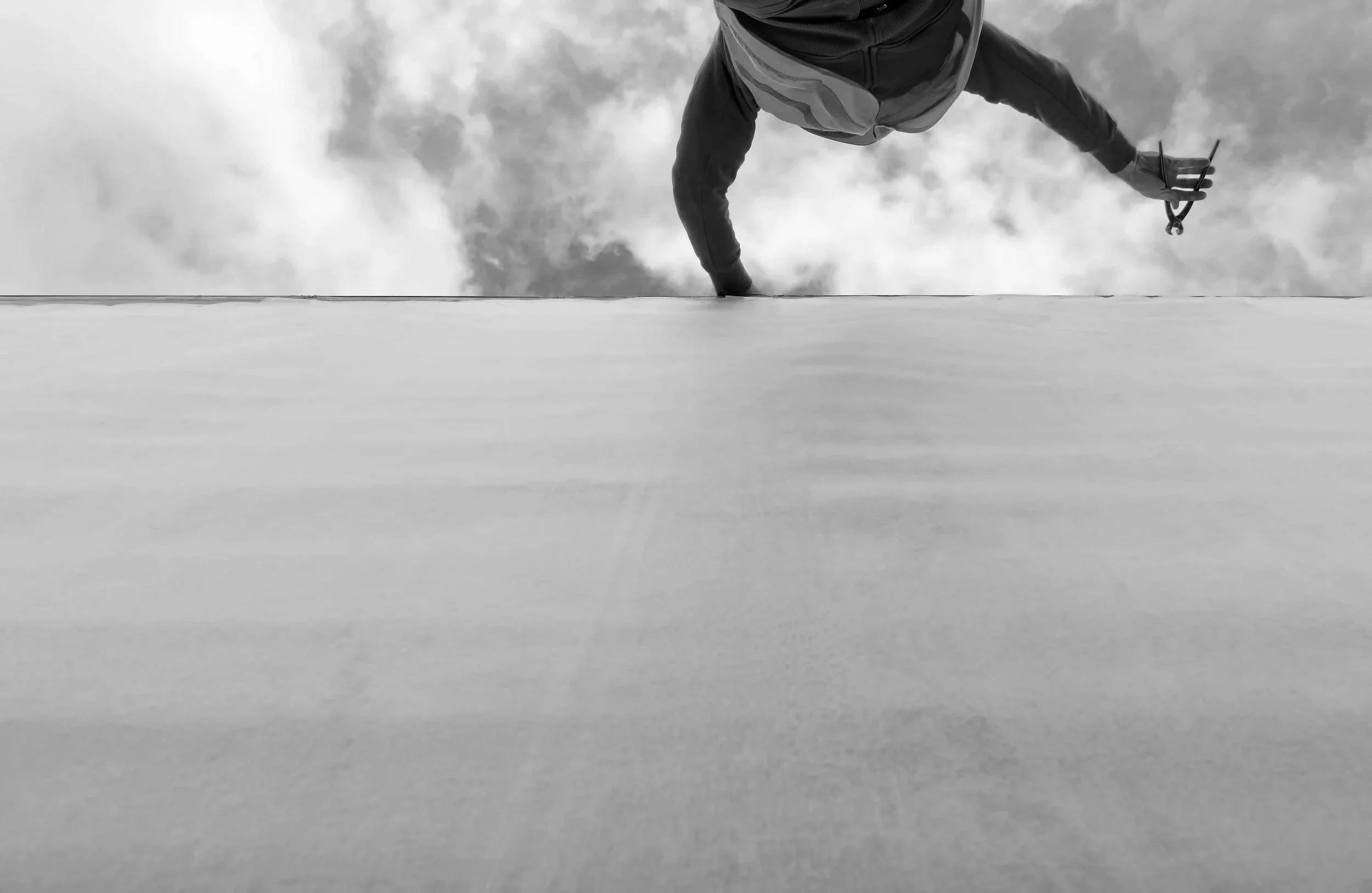 A person is climbing or hanging on a ledge or edge, captured from below, with a cloudy sky in the background. They are holding a smartphone or similar device in their hand.