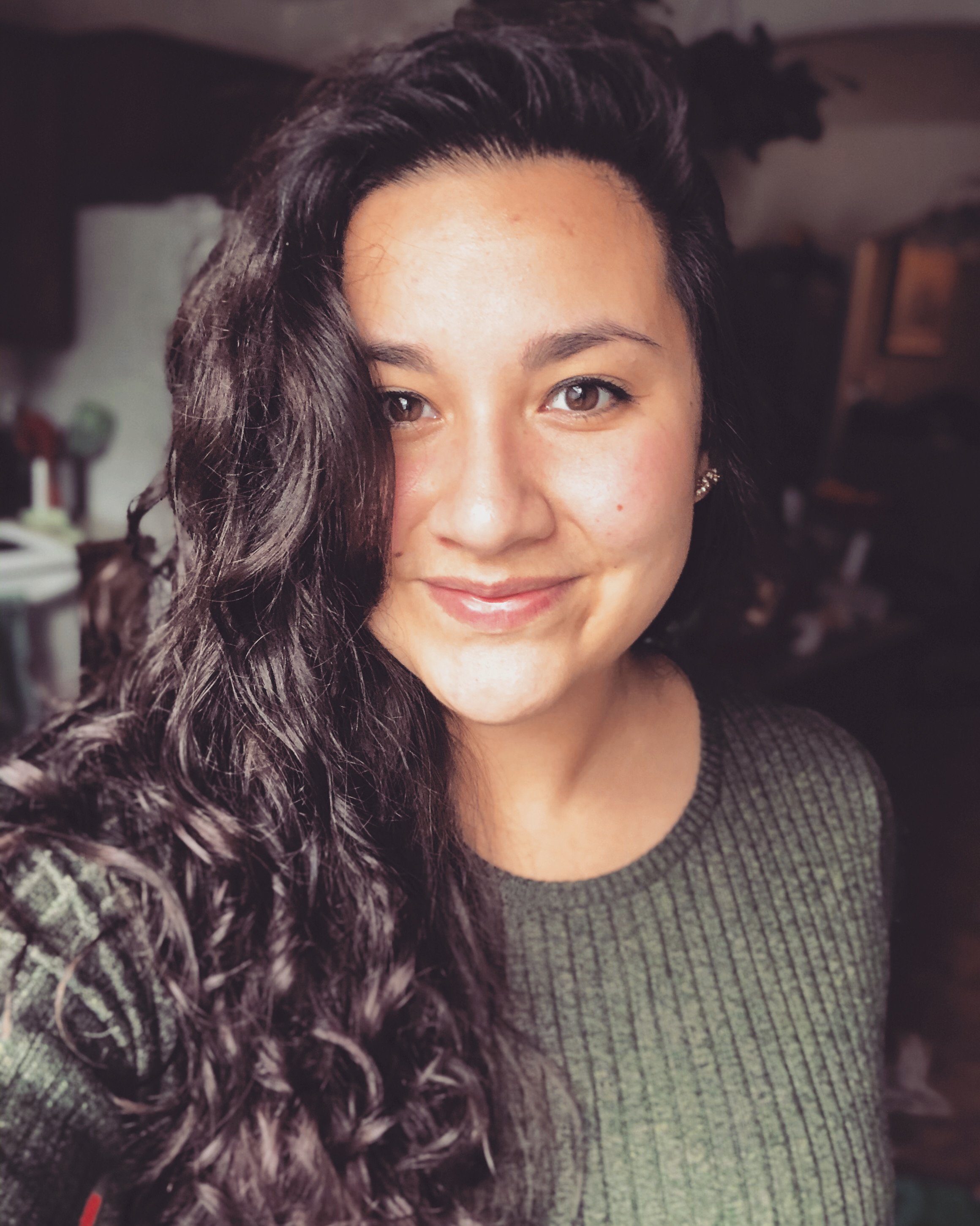 Close-up photo of a woman with long, dark, curly hair and light brown eyes, smiling gently, wearing a gray knit sweater.