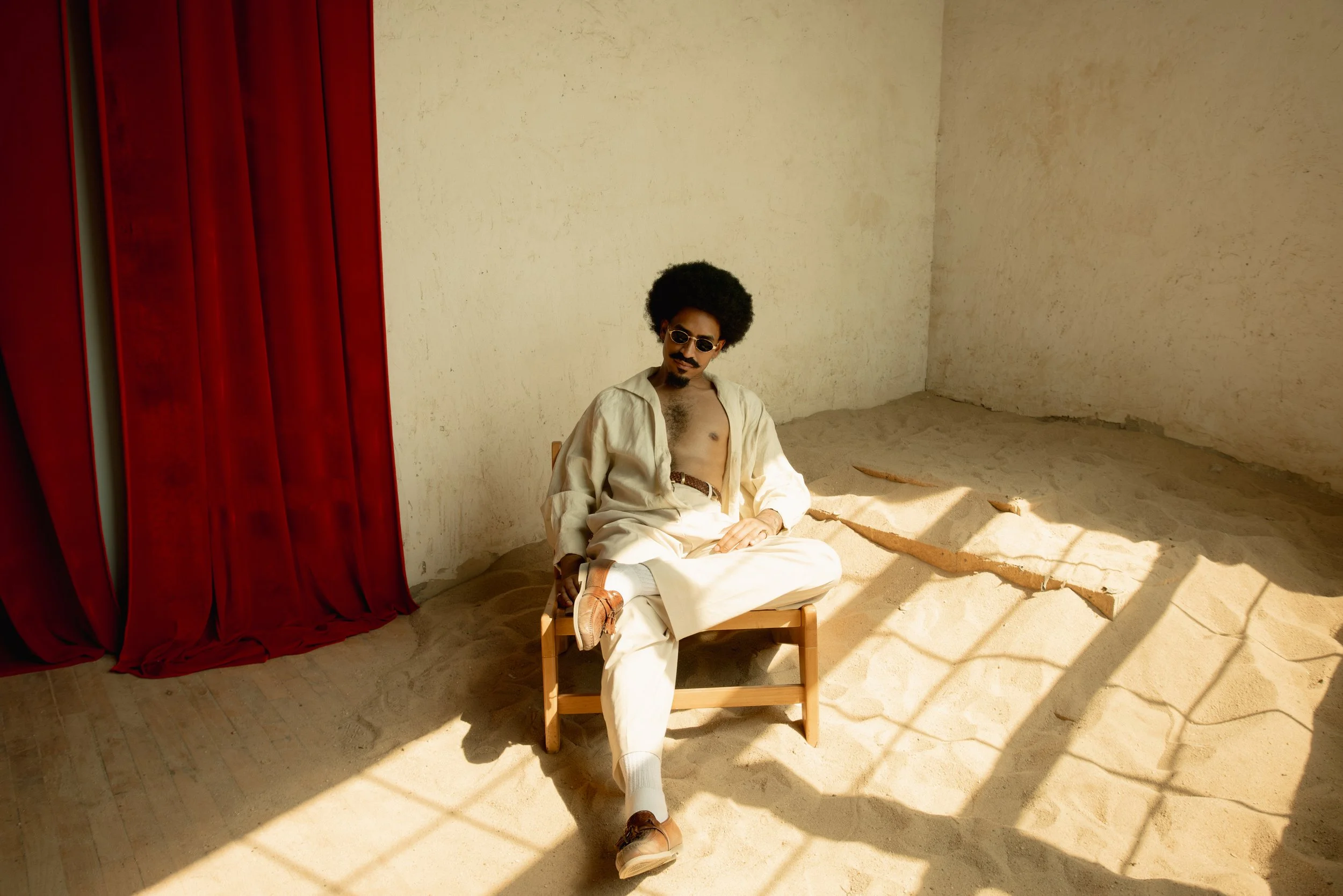 A man with curly hair, sunglasses, and a mustache is sitting on a small wooden chair in a room with a sandy floor and plain beige walls. He is dressed in white clothing and wearing brown shoes. A red curtain is partially visible on the left side. The