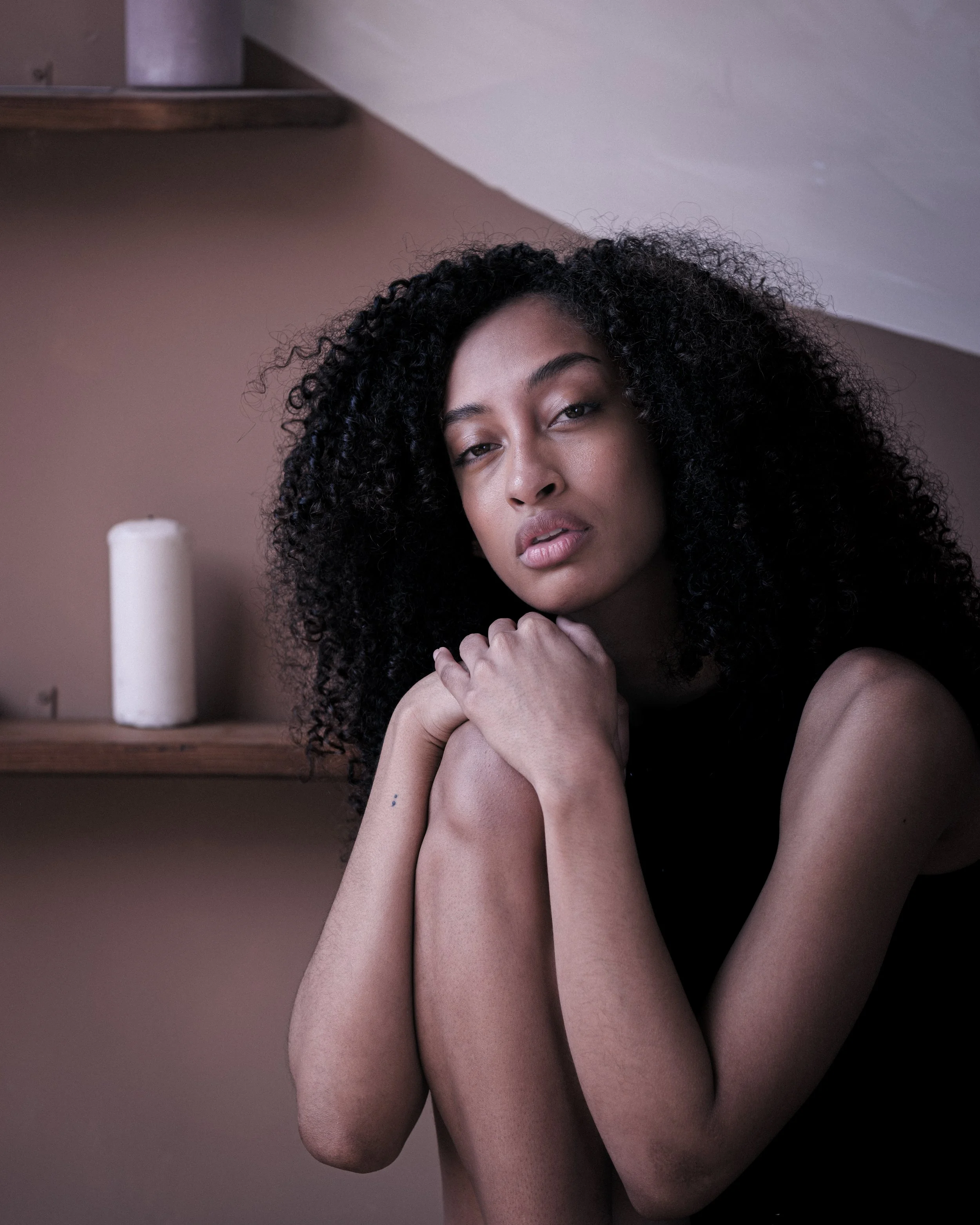 Close-up of a woman with curly hair, resting her chin on her crossed arms, with a neutral expression, in a softly lit setting with a candle in the background.