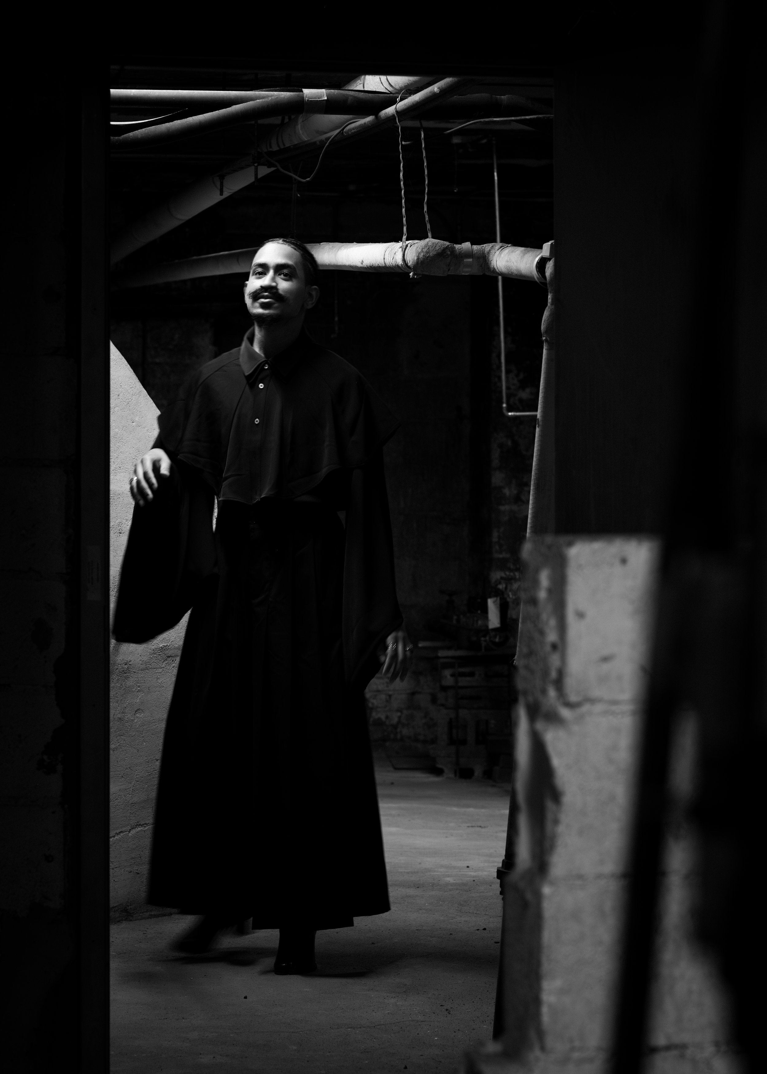A woman dressed in a dark gown and cape, standing in a dimly lit, industrial-looking space with pipes on the ceiling, as seen through a doorway or opening.