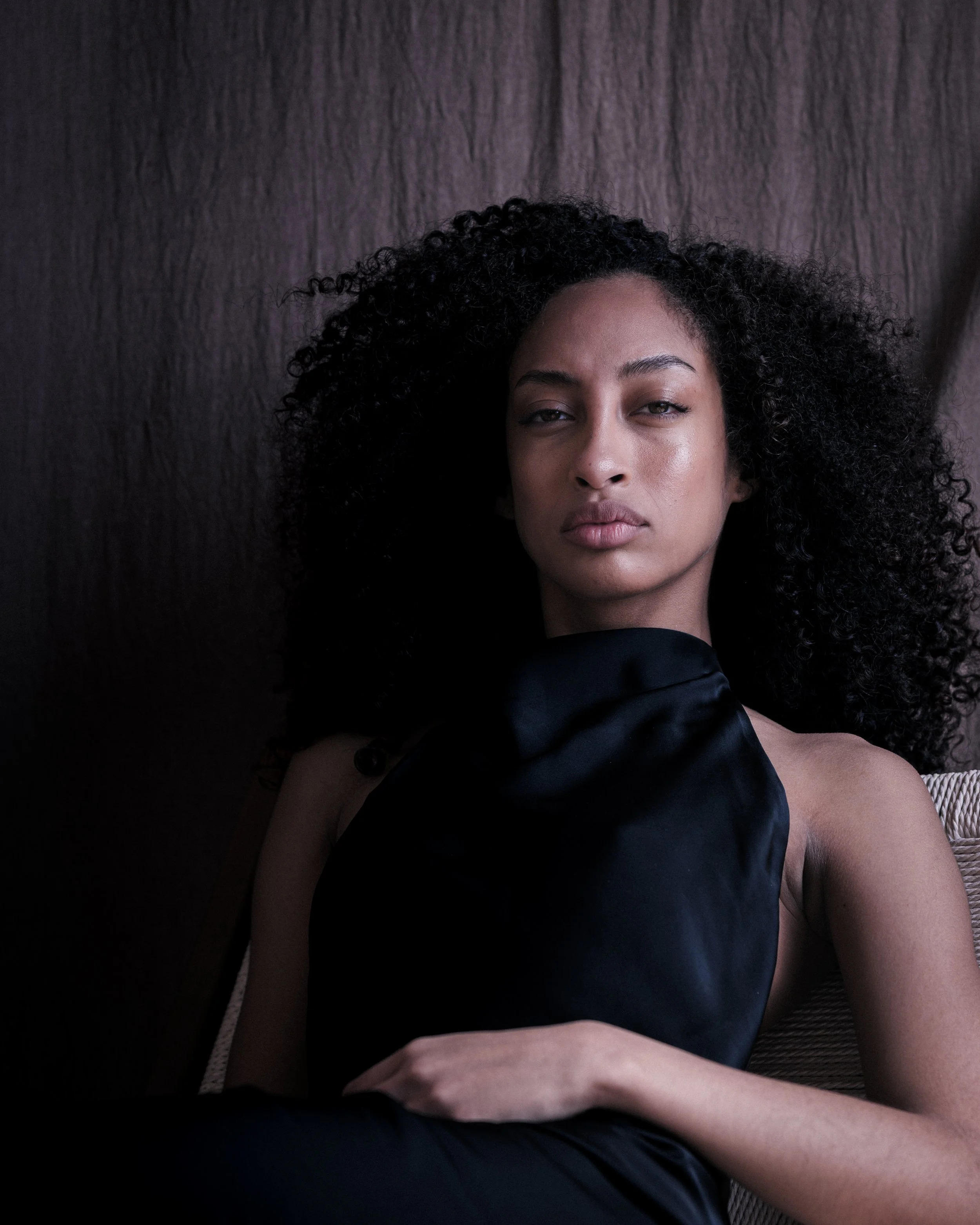 Close-up of a woman with curly black hair lying down on a dark textured surface, looking directly at the camera with a neutral expression, wearing a black sleeveless top.