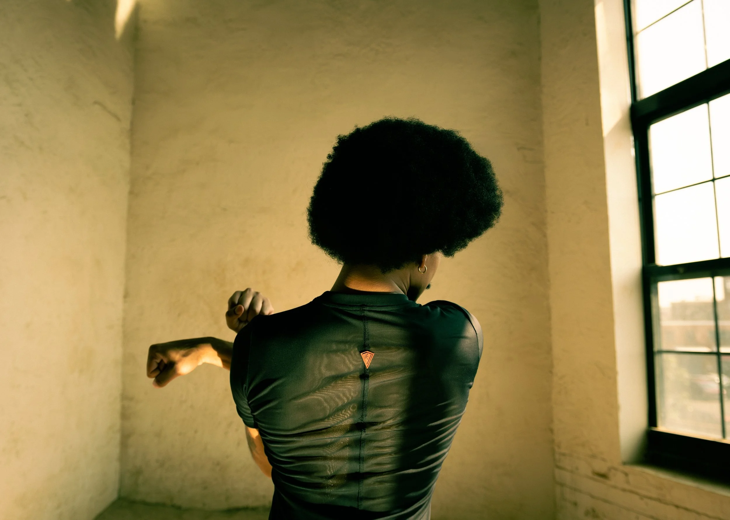 Back view of a person with an afro hairstyle wearing a black athletic shirt, standing indoors near a large window with sunlight shining through, with a plain wall behind.
