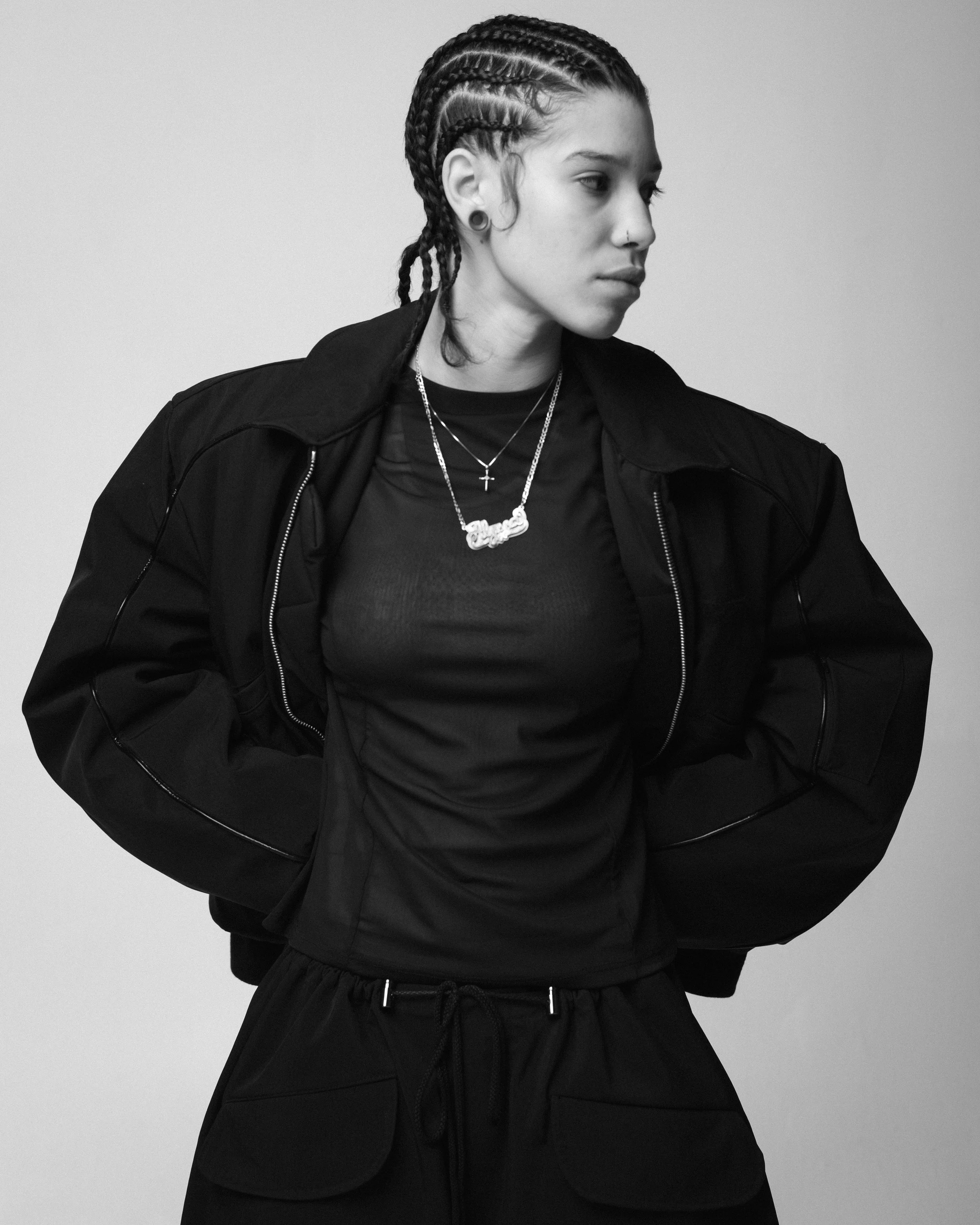 Black and white photo of a woman with braided hair, wearing a black jacket, layered necklaces, and a nose ring, standing against a plain background.