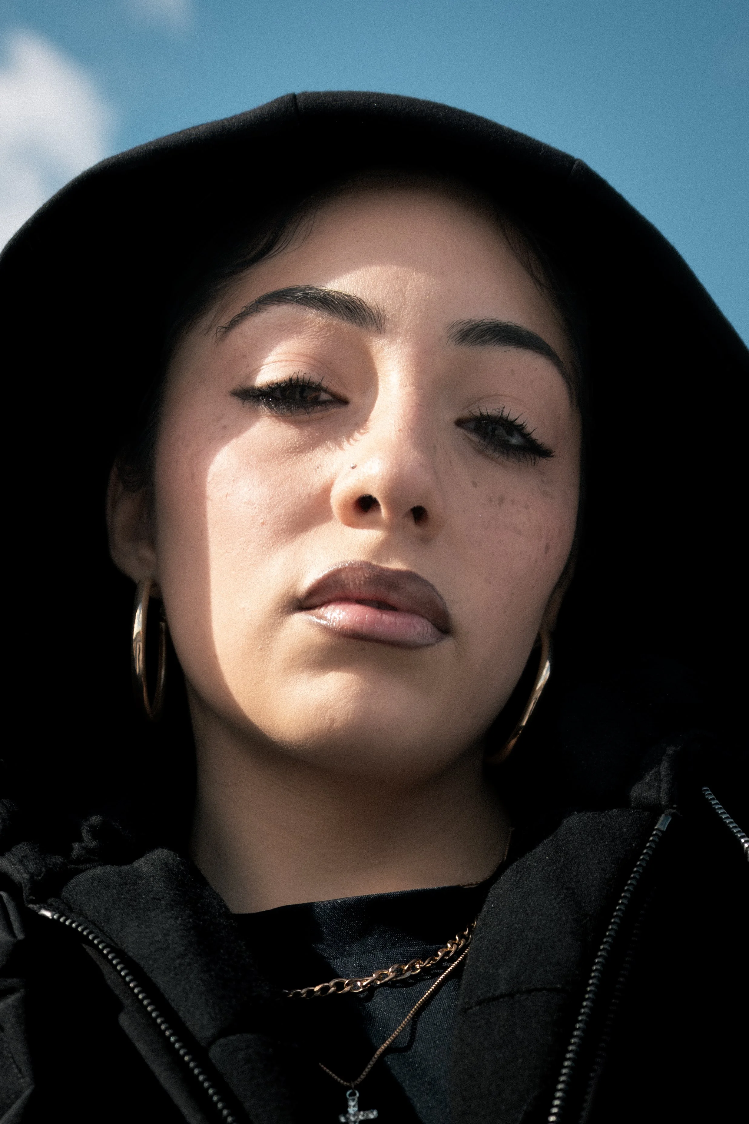 Close-up of a young woman with dark hair and pierced ears, wearing a black hoodie with the hood up, gold hoop earrings, and layered necklaces, outdoors with a blue sky in the background.
