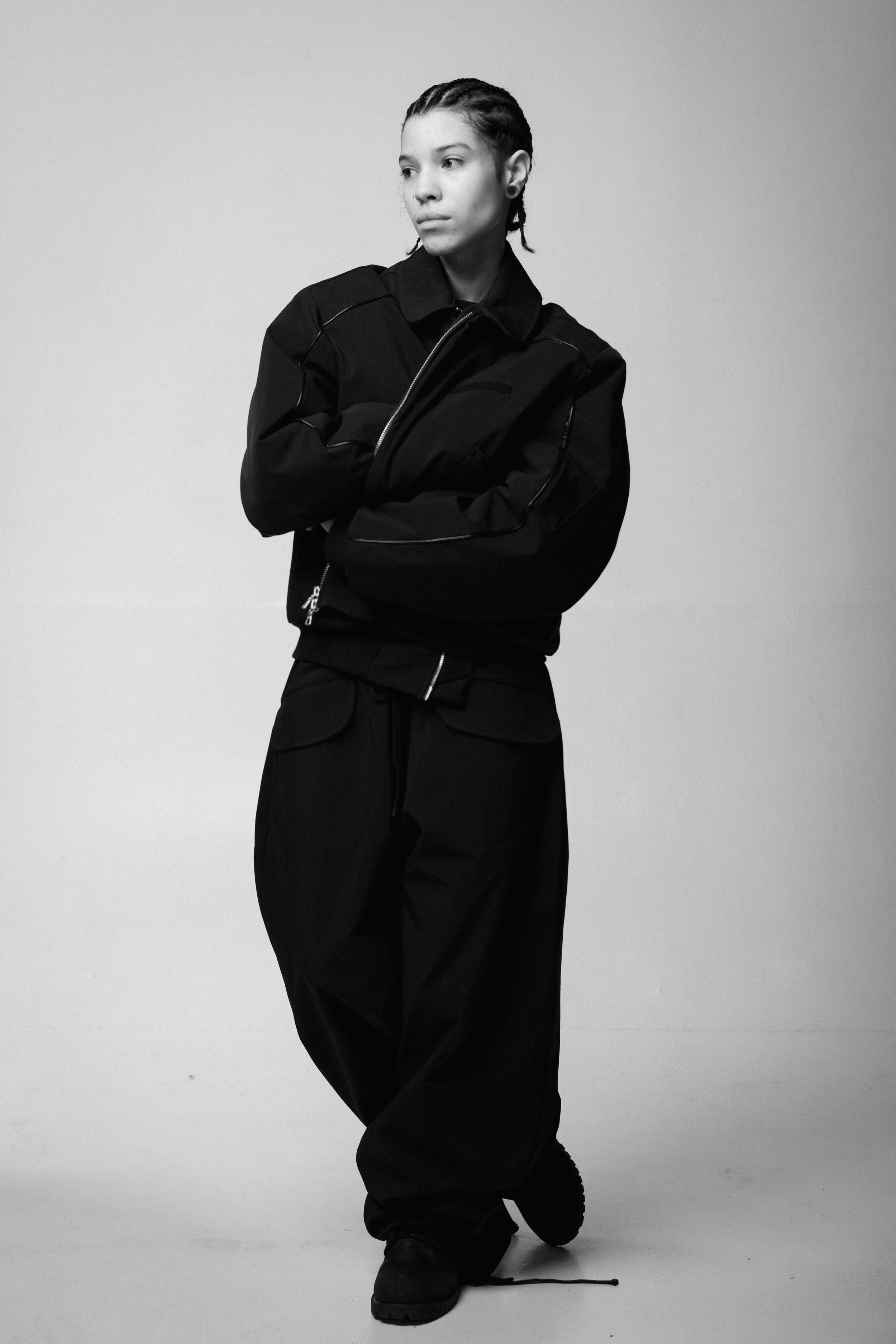 Black and white photo of a person with braided hair, wearing a black jacket and black pants, kneeling on one knee with crossed arms, in front of a plain light background.