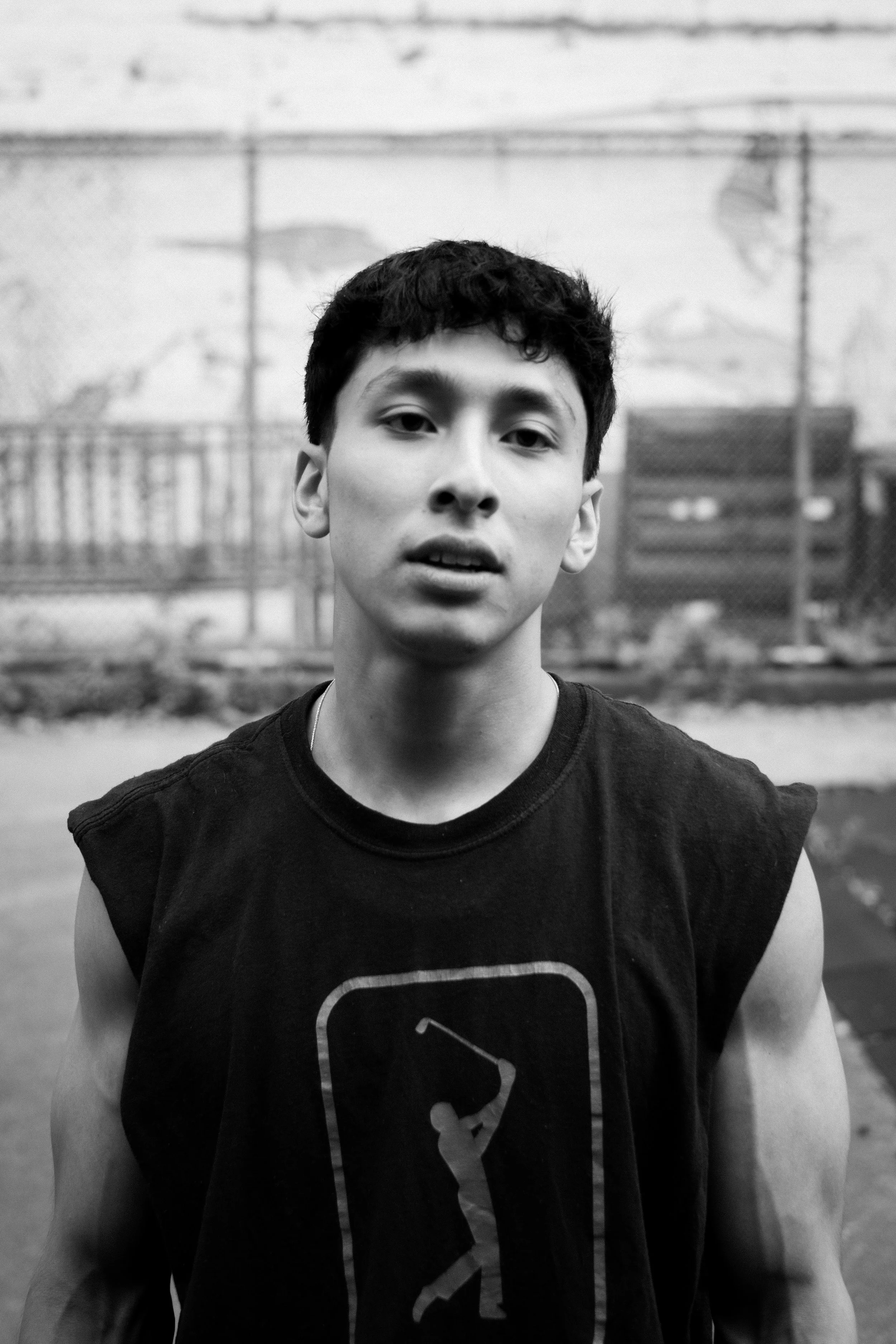 Black and white photo of a young man with short dark hair wearing a sleeveless shirt with a graphic design, standing outdoors with a chain-link fence and some trees in the background.