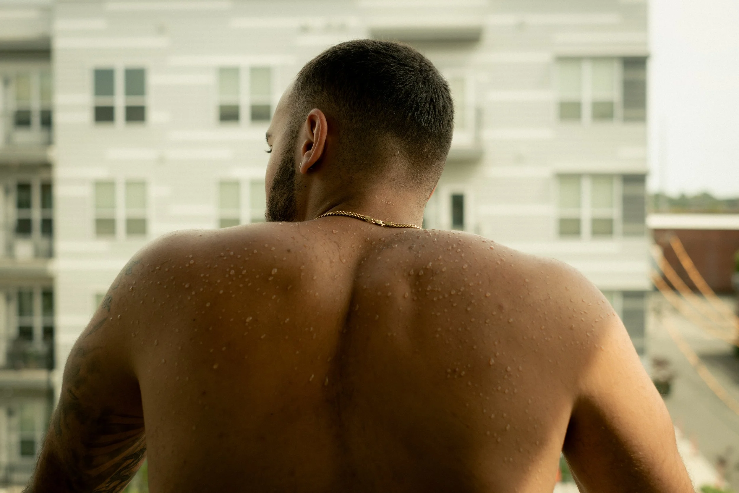 A man with short dark hair and a beard, seen from behind, is shirtless and on a balcony or terrace. Water droplets are visible on his back and shoulder. In the background, there are multistory residential buildings.