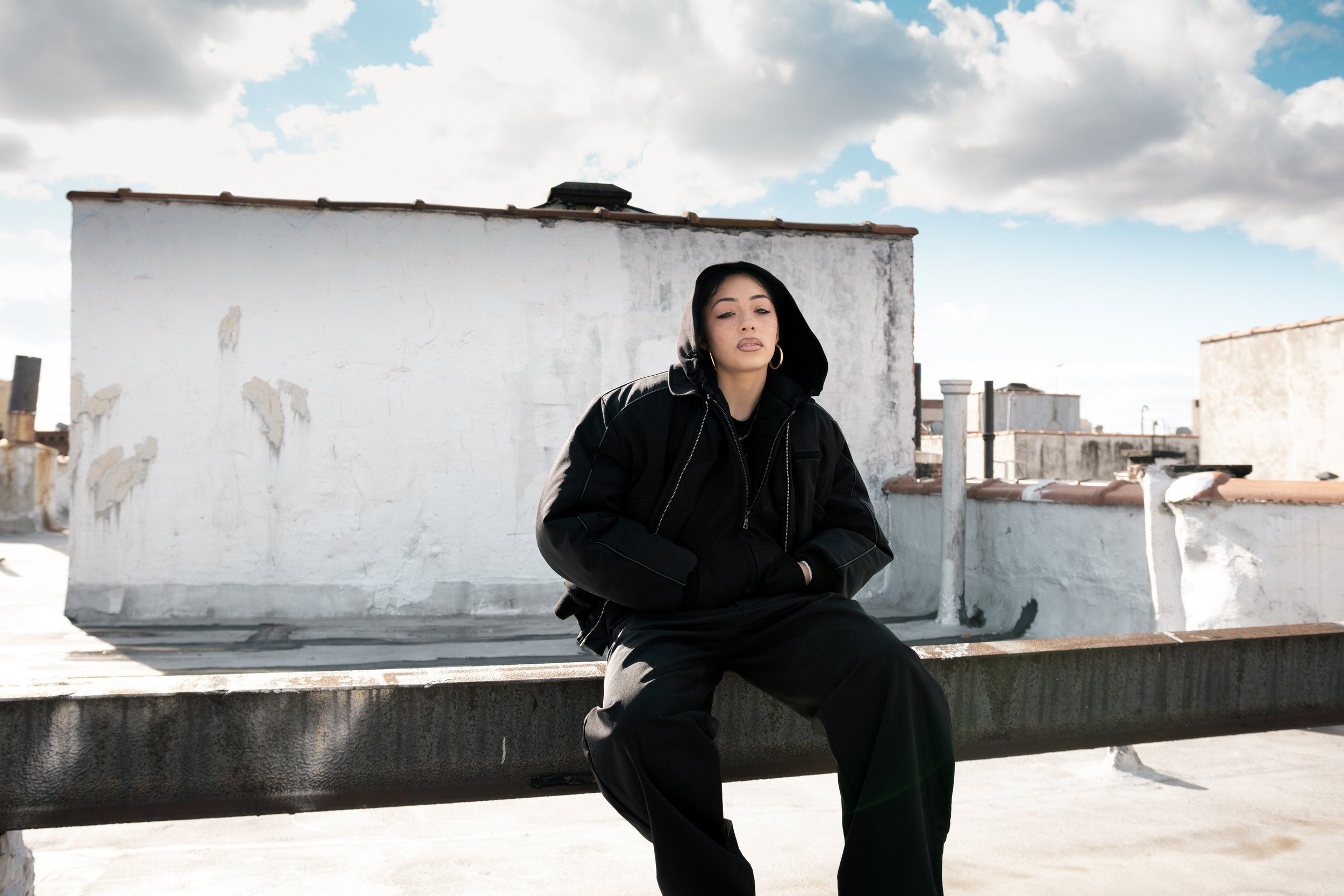 Young woman with black hair, wearing a black hoodie and large hoop earrings, sitting on a rooftop with white walls and cloudy sky in the background.
