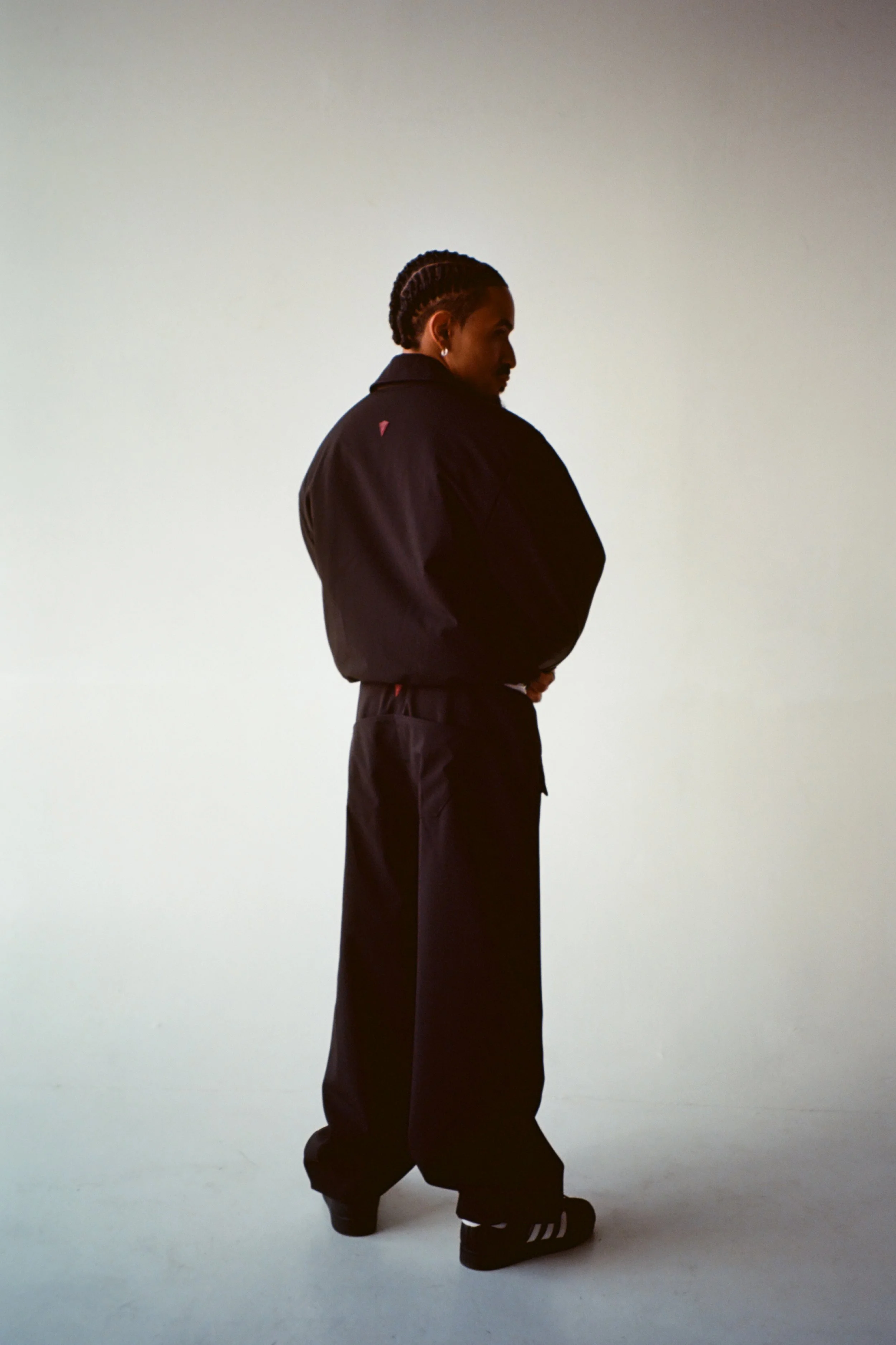 Side profile of a person walking, dressed in black pants, black jacket, and black sneakers with white stripes, standing against a plain white background.