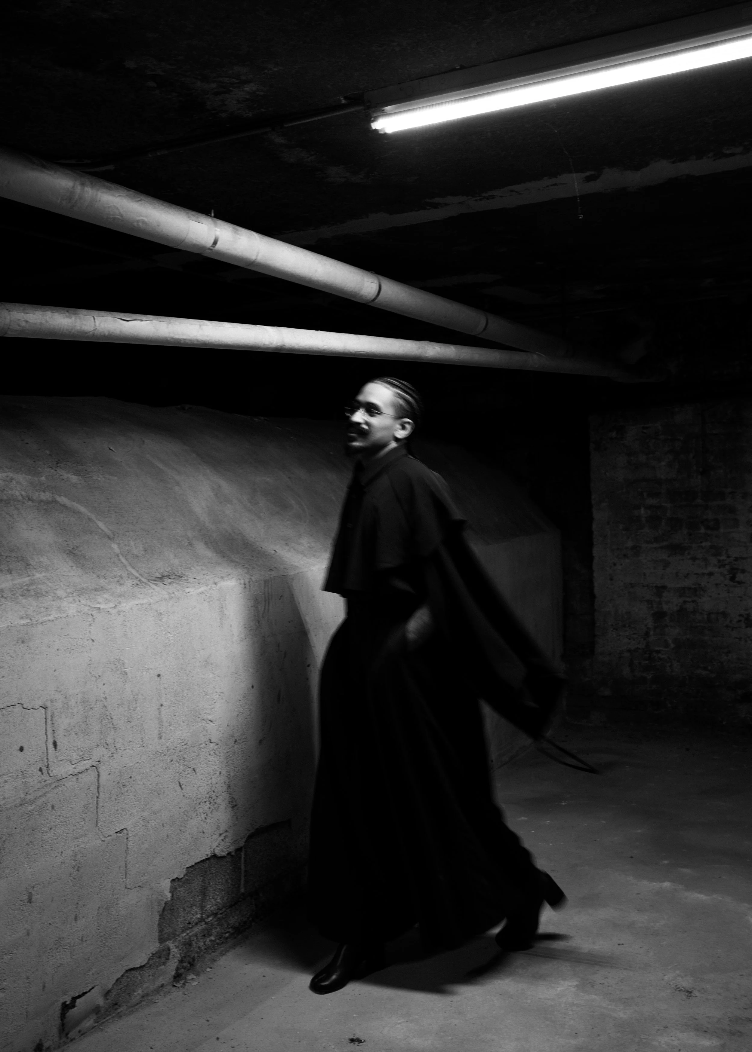 A person with short hair and glasses dressed in dark clothing walking in a dimly lit underground parking garage with concrete walls, pipes, and overhead fluorescent lighting.