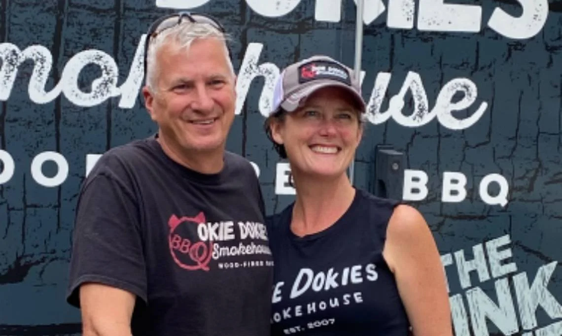 A smiling man and woman wearing black T-shirts with the logo of 'Okie Dokie's Smokehouse' standing in front of a sign for the barbecue restaurant.