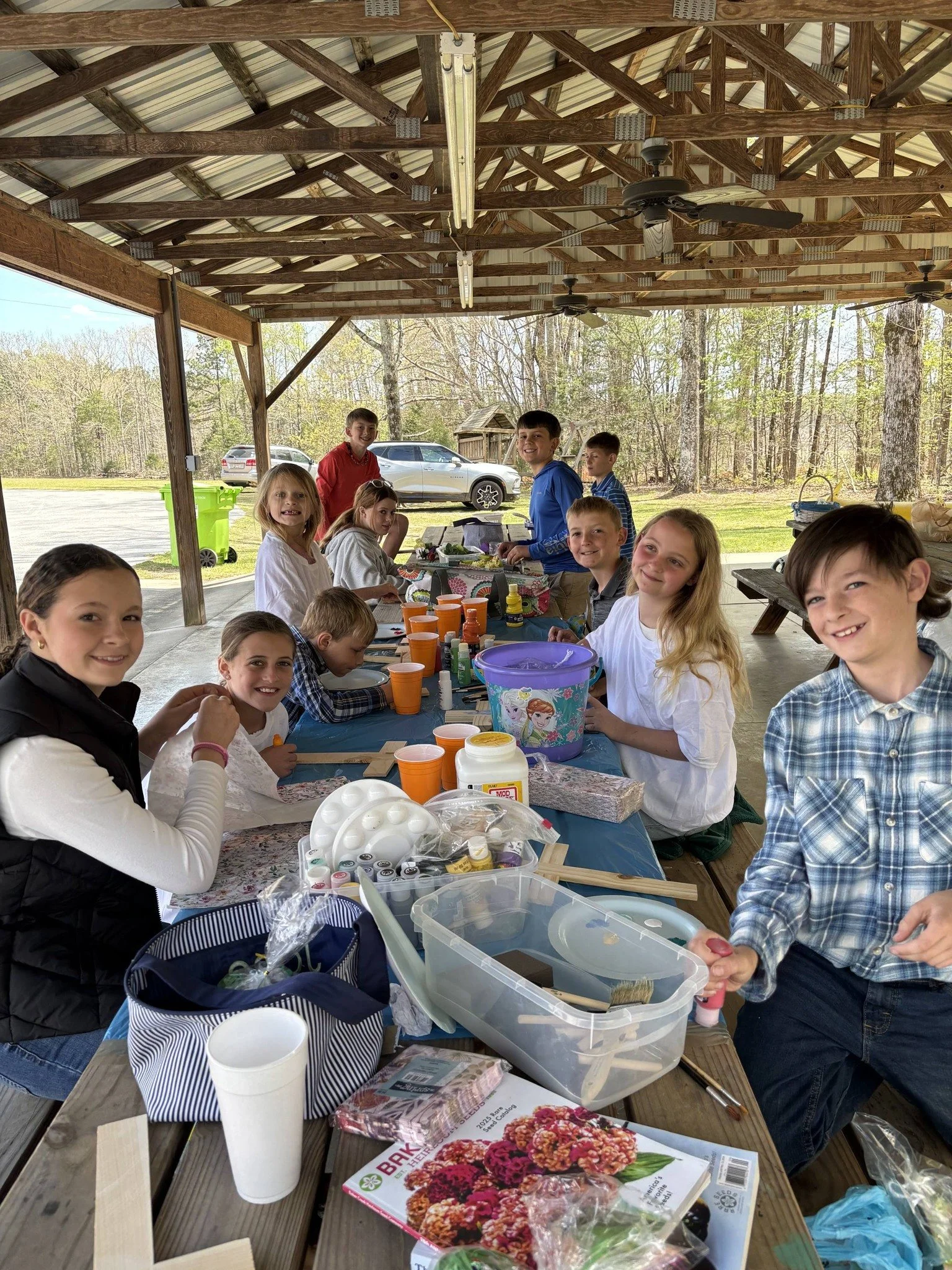 Group of children happily gathered at a table under a wooden pavilion, engaging in arts and crafts activities with various supplies, with a wooded outdoor background and cars parked outside.