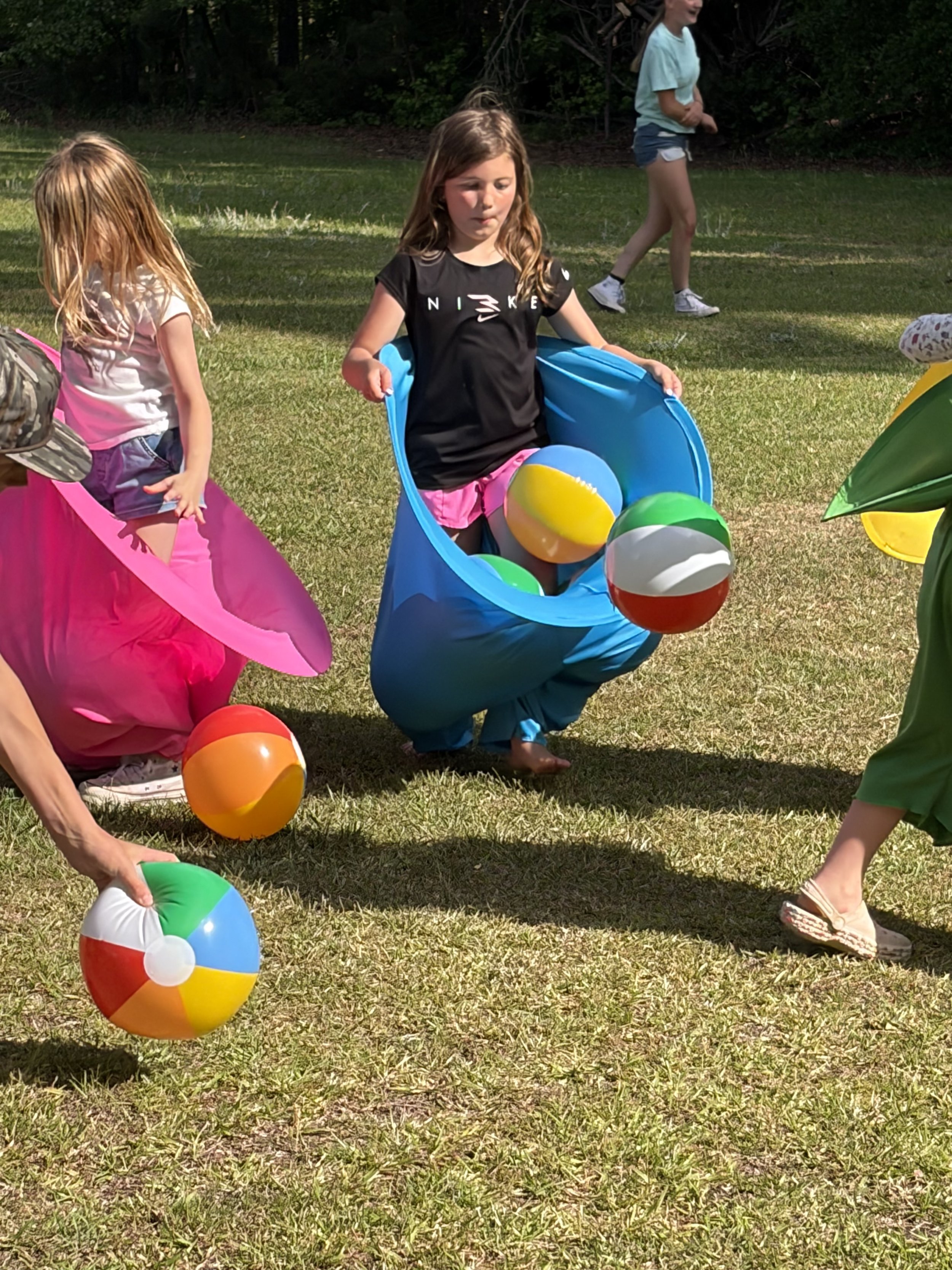 Children playing outdoors with beach balls, some entering colorful fabric tunnels on grassy ground, in a park or backyard setting with trees in the background.