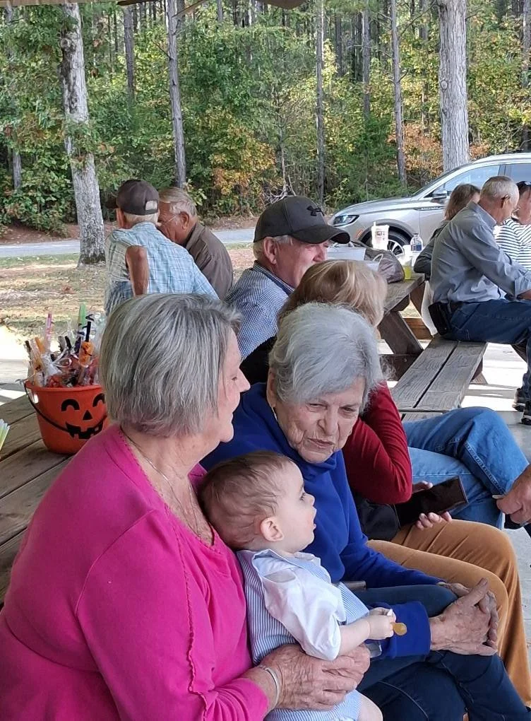 Group of people sitting on a wooden picnic table outdoors, with a background of trees and a car. An elderly woman holding a toddler, both facing a woman in a pink top.