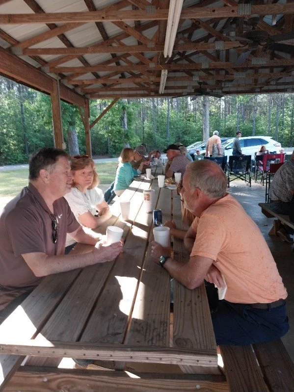 People sitting at picnic tables under a wooden shelter, drinking and socializing outdoors on a sunny day.