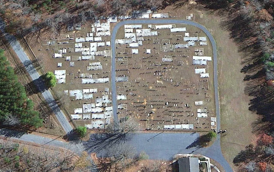 Aerial view of a cemetery with uneven terrain, rows of tombstones, and surrounding trees.