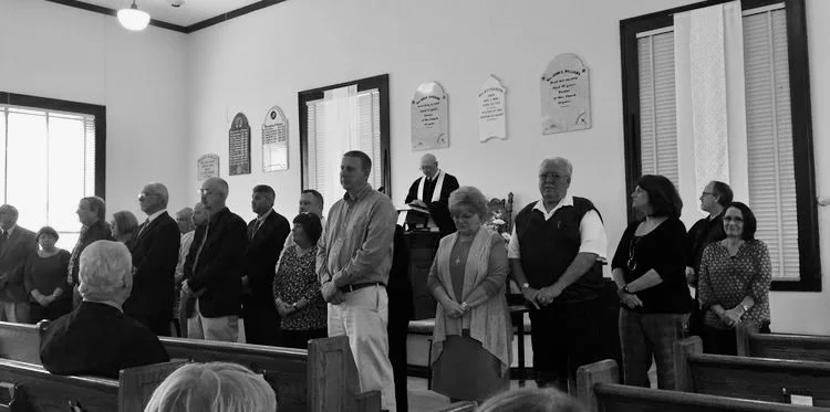 Group of people standing in a church during a service or ceremony, with some seated in pews and others standing at the front, with plaques or memorials on the wall behind.