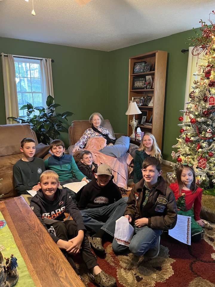 A group of children and an elderly woman gathered in a living room decorated for Christmas, with a Christmas tree on the right side and a green-painted wall with a bookshelf on the back wall.