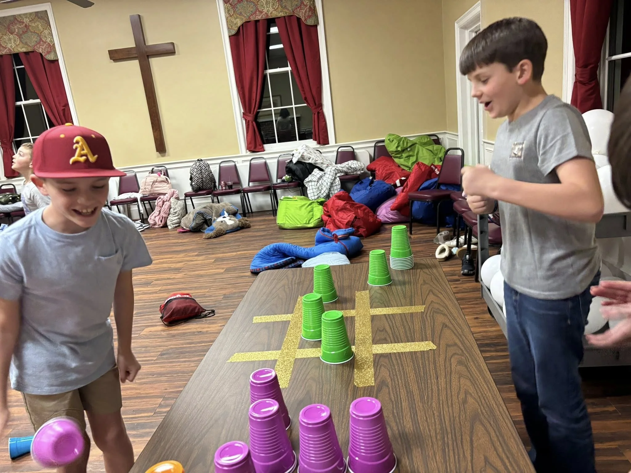 Children playing a stacking game with colored cups in a room with red curtains, a large cross on the wall, and chairs with backpacks and bags on them.