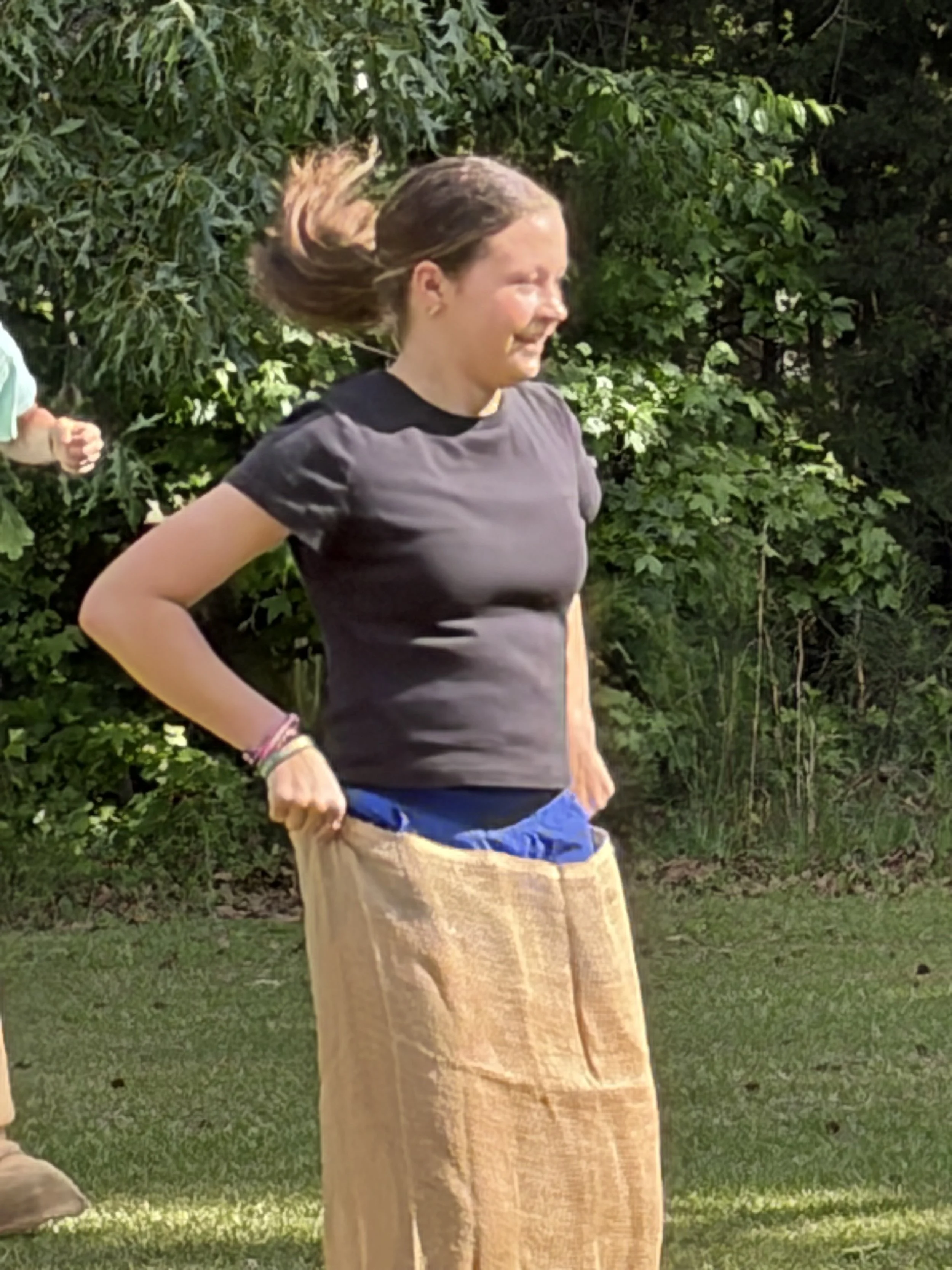 A young woman with brown hair in a ponytail is outdoors in a grassy area surrounded by green foliage. She is wearing a dark t-shirt and has multiple bracelets on her left wrist. She is participating in a sack race, with her hands holding a tan burlap sack around her waist.