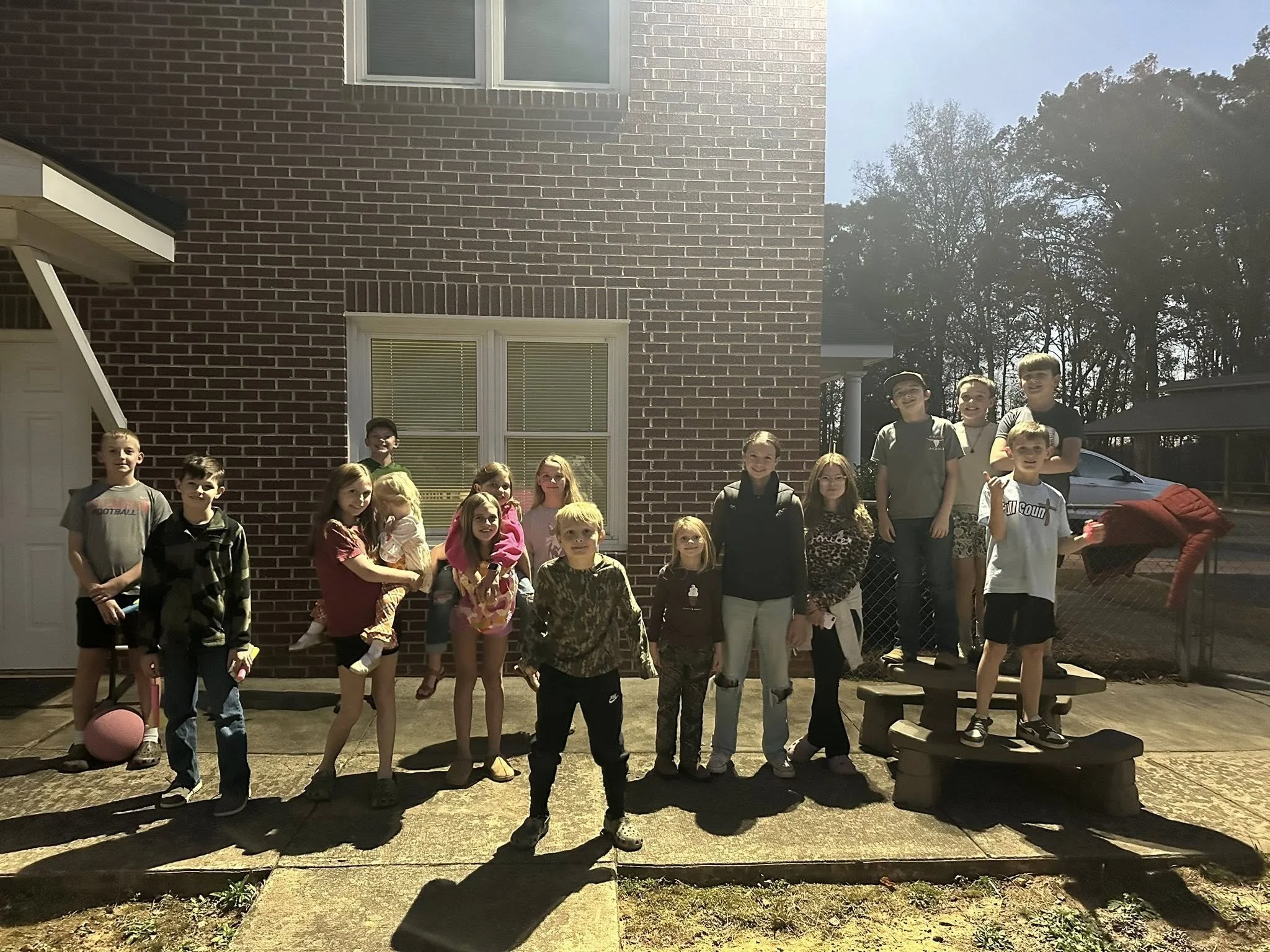 Group of children and teenagers standing outside on a porch in front of a brick house with multiple windows and trees in the background, illuminated by sunlight.