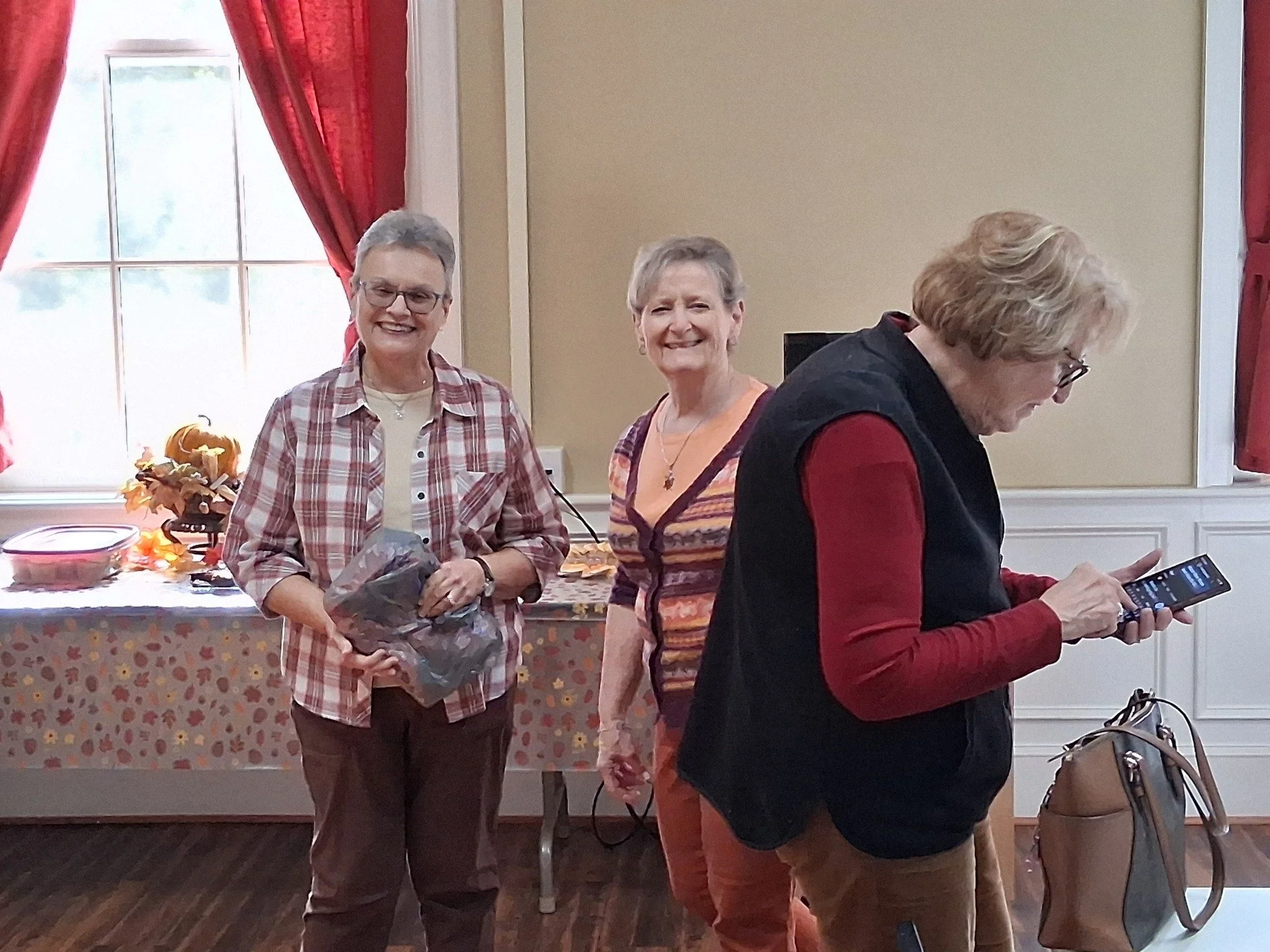 Three women are standing in a room with red curtains and a decorated table in the background. The woman on the left is smiling, wearing glasses, a plaid shirt, and holding a bouquet. The woman in the middle is smiling, wearing a colorful top. The woman on the right is looking down at her phone, wearing glasses, a black vest over a red shirt, and brown pants.