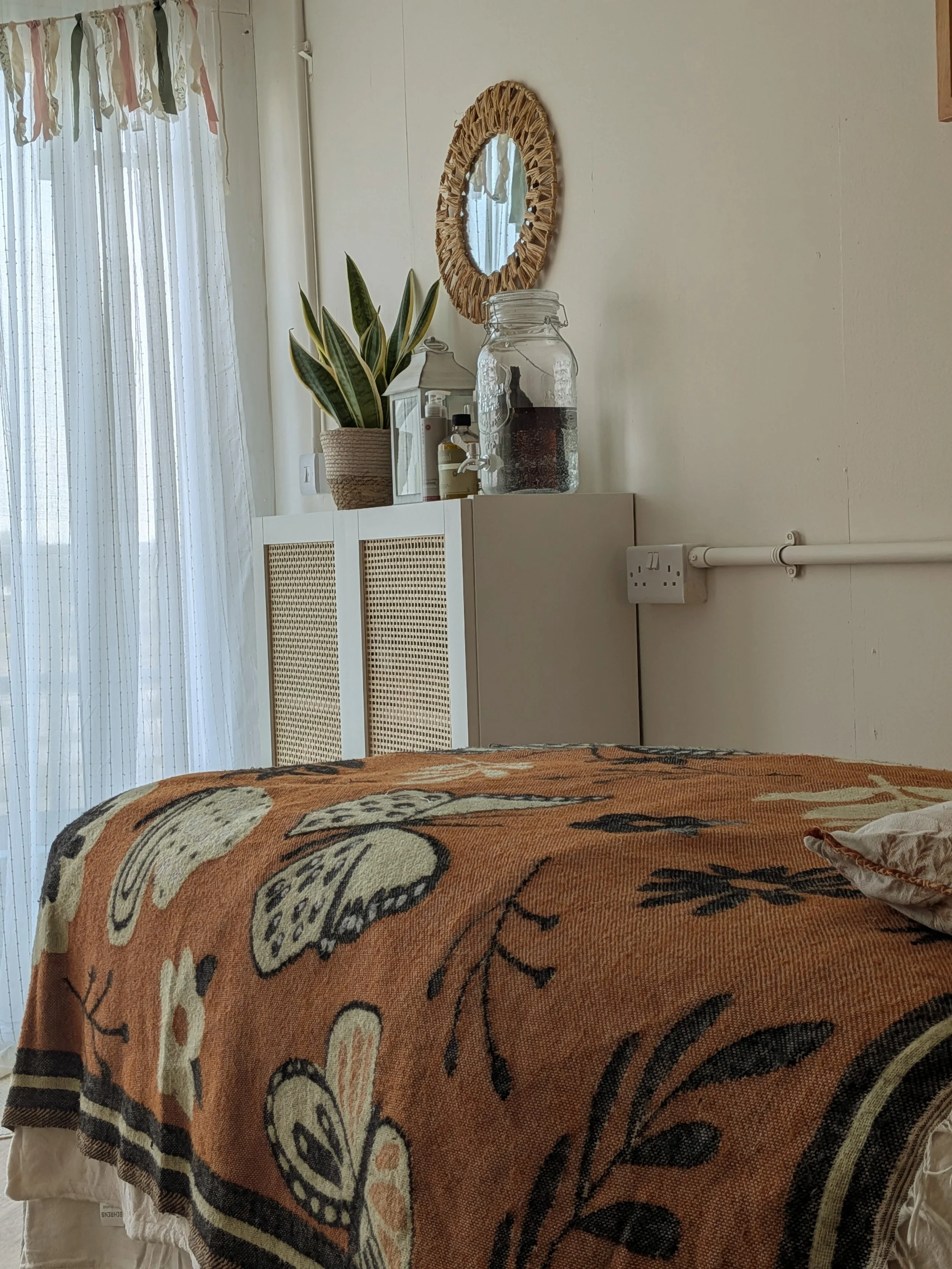 A warm and welcoming massage therapy with a massage couch covered by an orange blanket with a floral pattern. Behind the couch, there is a white cabinet and a glass jar on top.