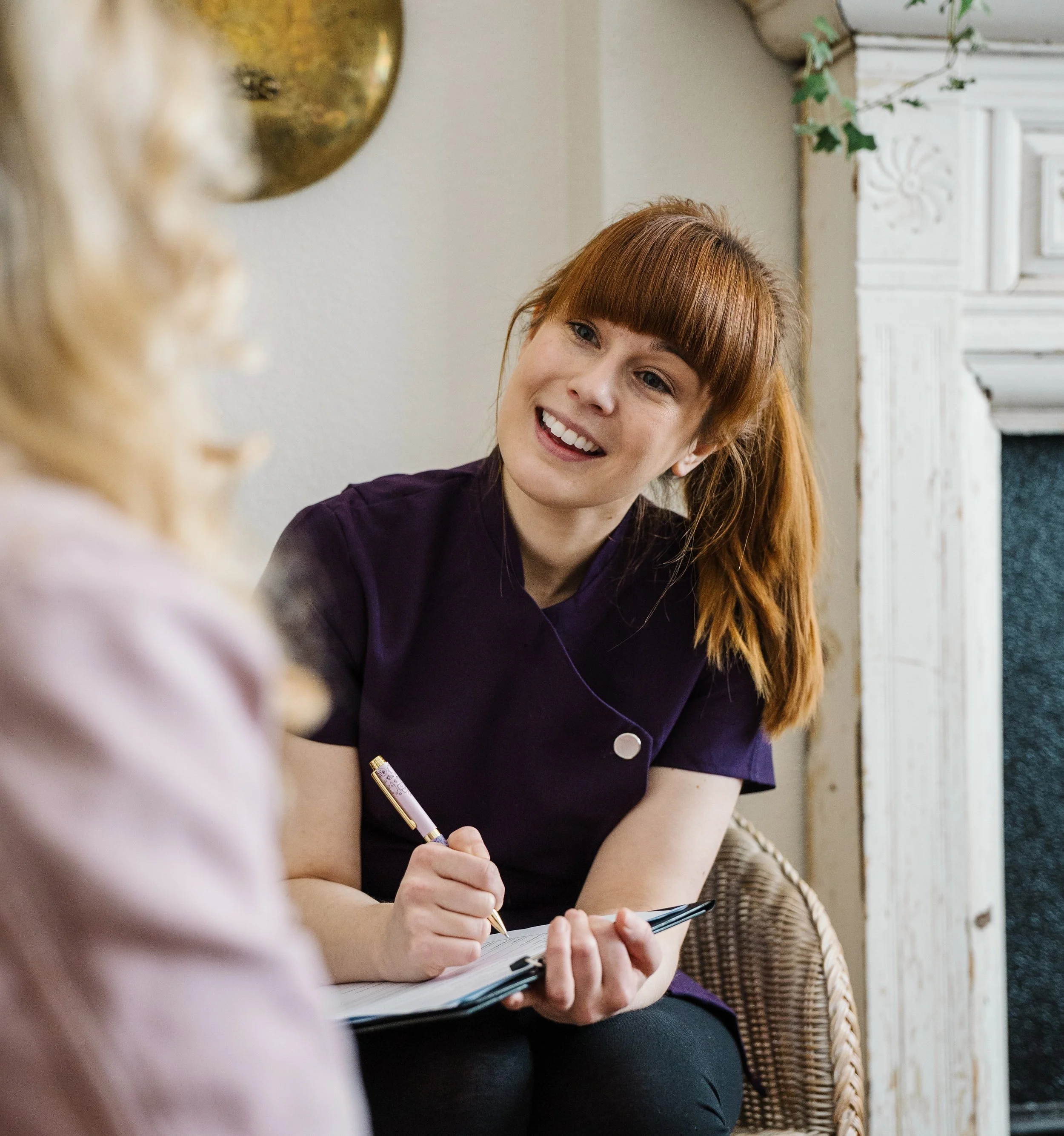 Tansy smiling and taking notes, while a person with blonde hair is in the foreground out of focus. The background shows a white fireplace and a plant with green leaves.