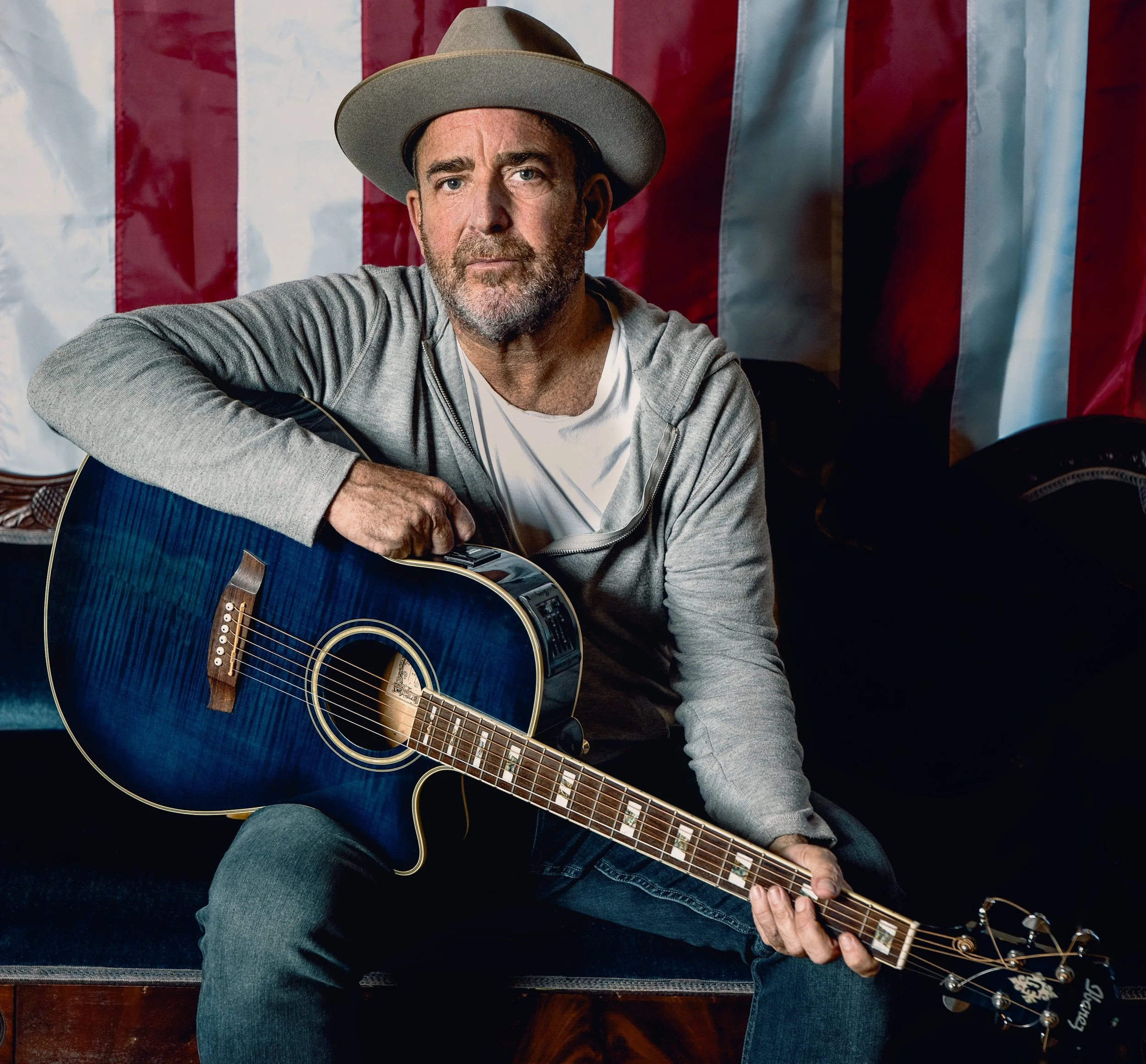 A middle-aged man with a beard and wearing a gray fedora hat and gray hoodie, sitting with an acoustic guitar in front of a backdrop of an American flag.