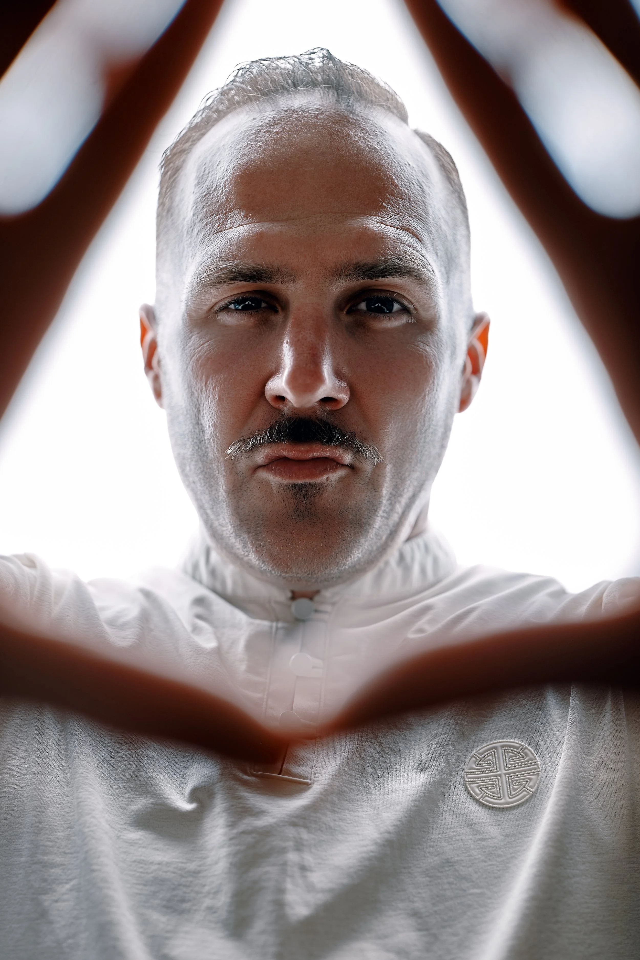Close-up selfie of a man, Front Row Frankie, with a mustache and slicked-back hair, wearing a white traditional Asian-inspired shirt with a circular embroidered symbol on the chest, framed by his hands forming a triangle shape.