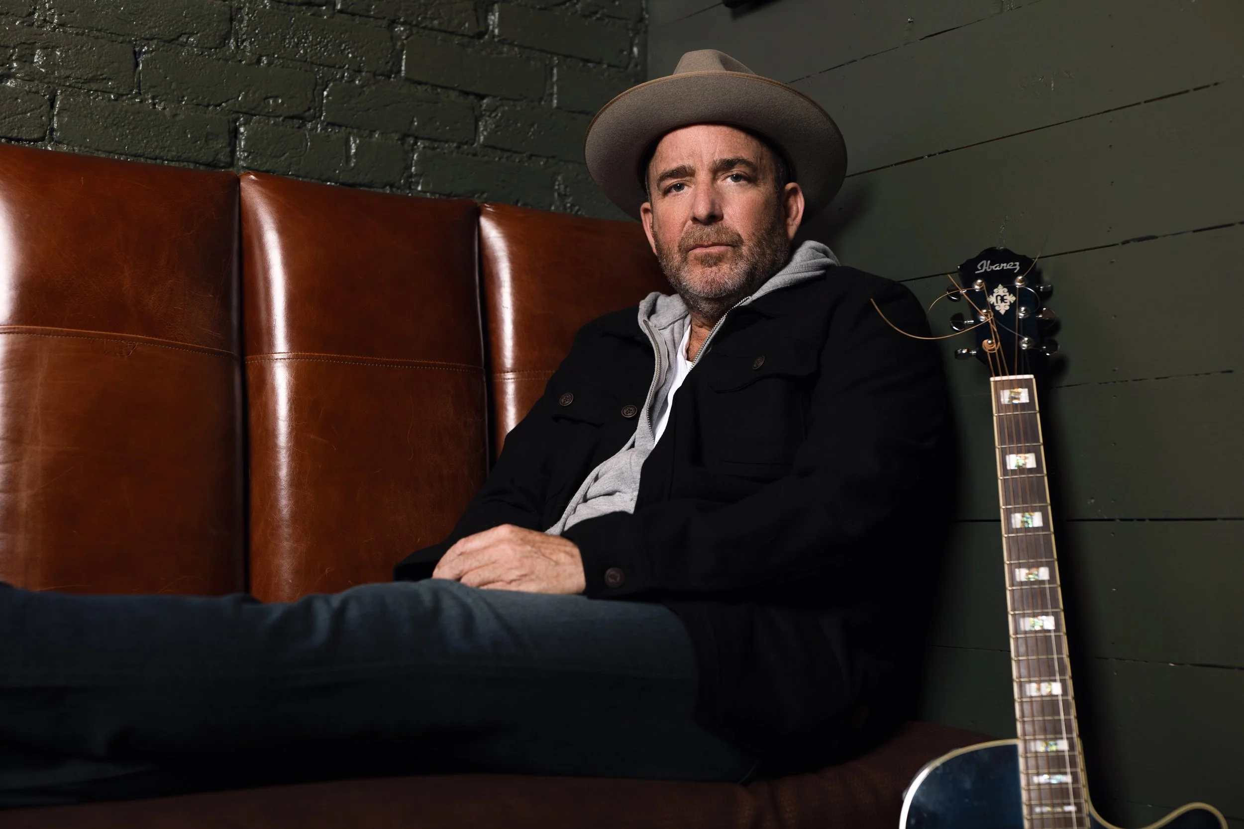 A man wearing a gray hat and a black jacket sitting on a brown leather booth next to a green wooden wall, with a guitar lying beside him.