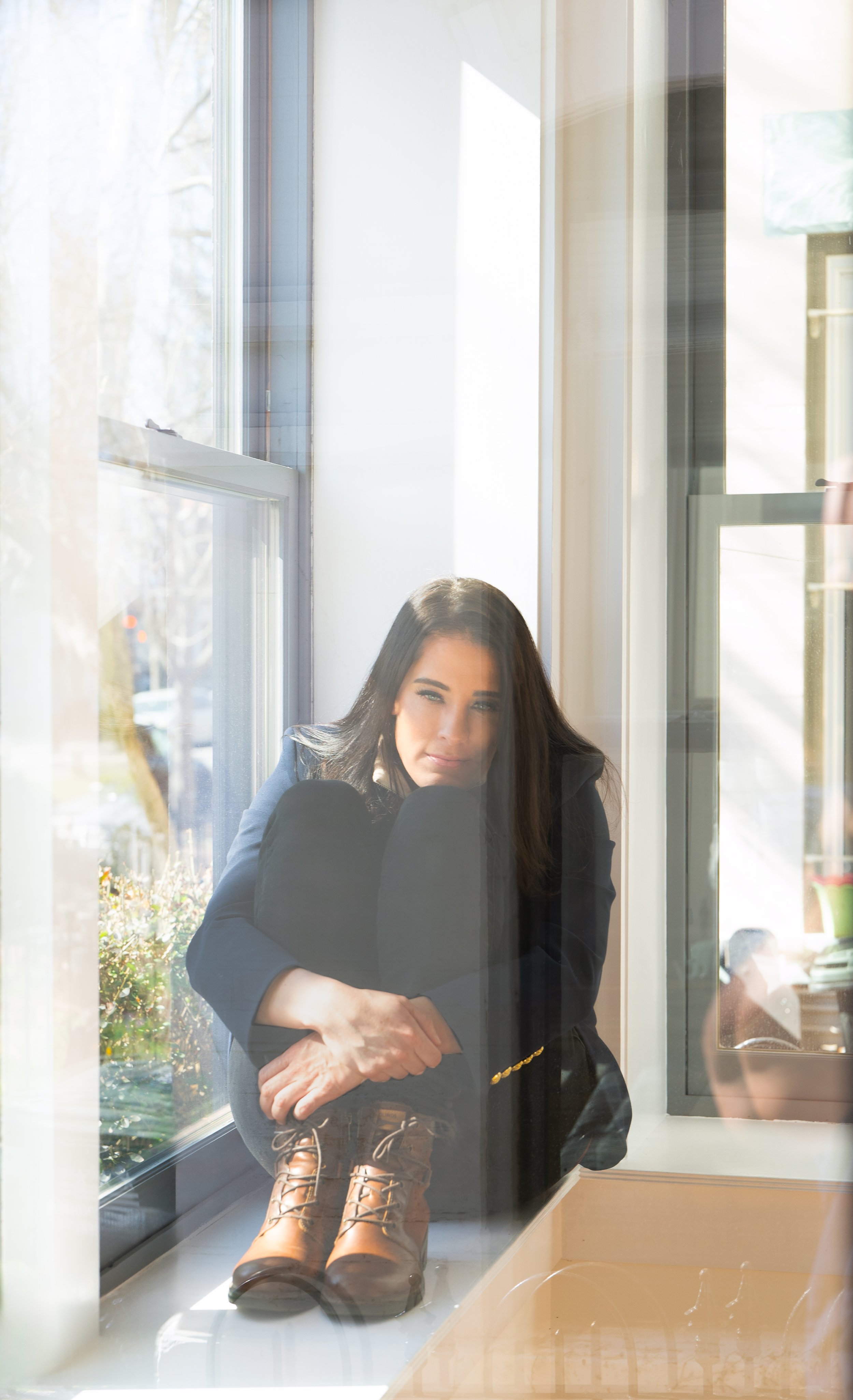 Young woman with long dark hair sitting on a window ledge, wearing a dark blazer, holding her knees with her arms, and looking through a window on a sunny day.