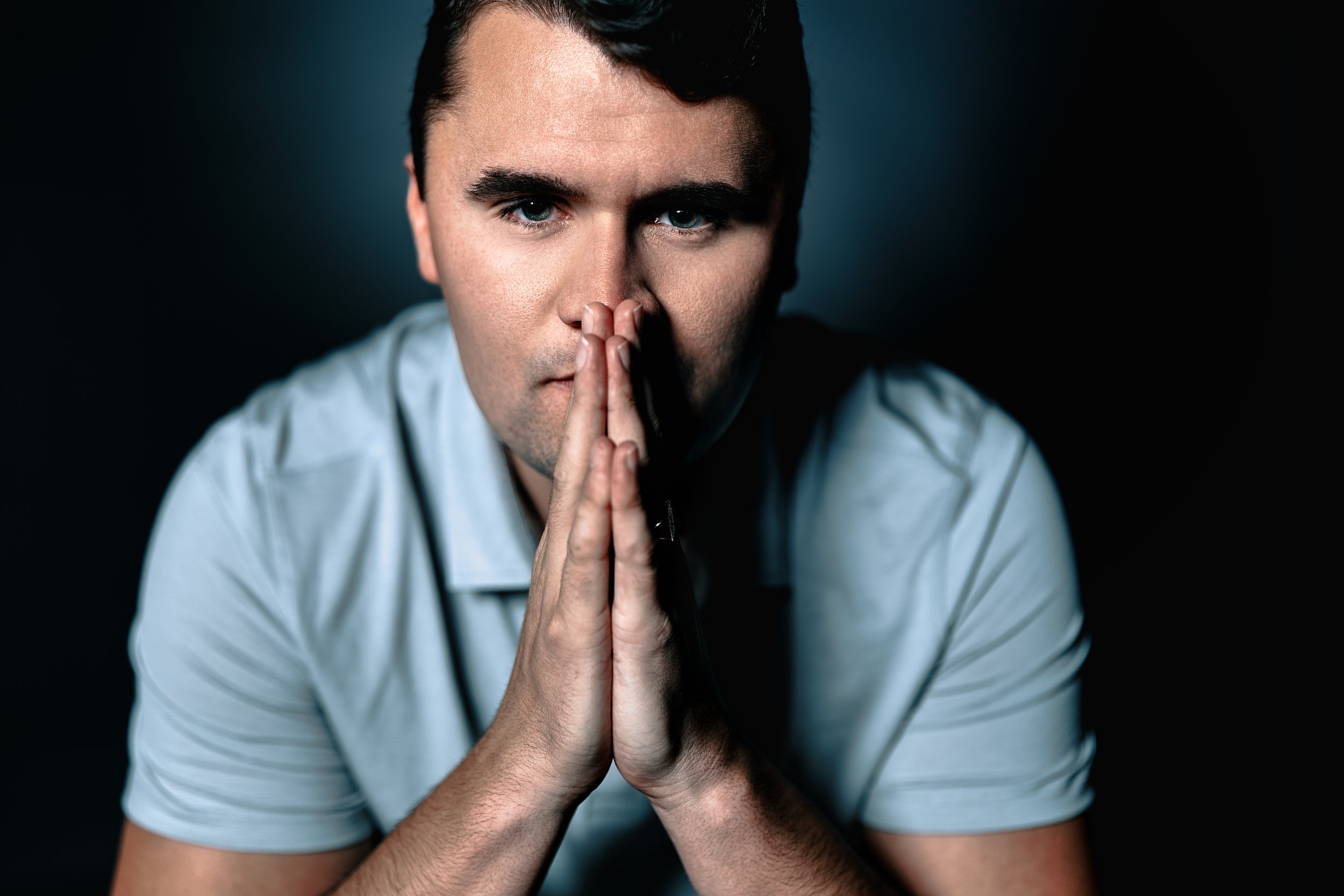 A young man with dark hair and blue eyes, wearing a light blue shirt, holds his hands together in a prayer-like gesture near his face, with a serious expression, against a dark background.