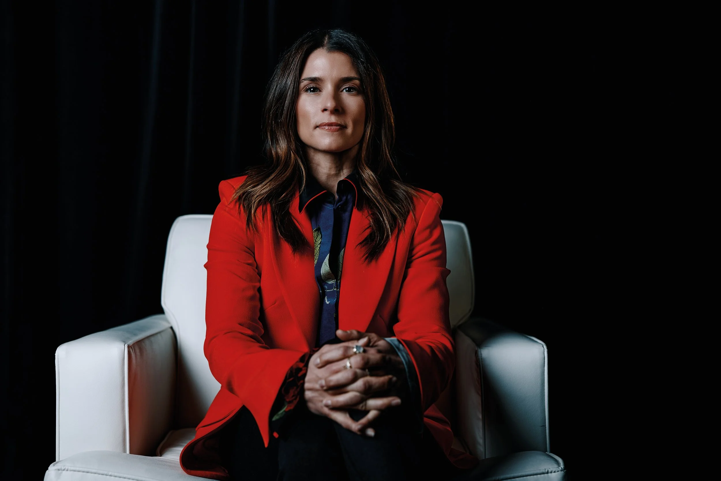 A woman with brown hair in loose waves, wearing a red blazer over a colorful blouse, sitting on a white chair against a black background.