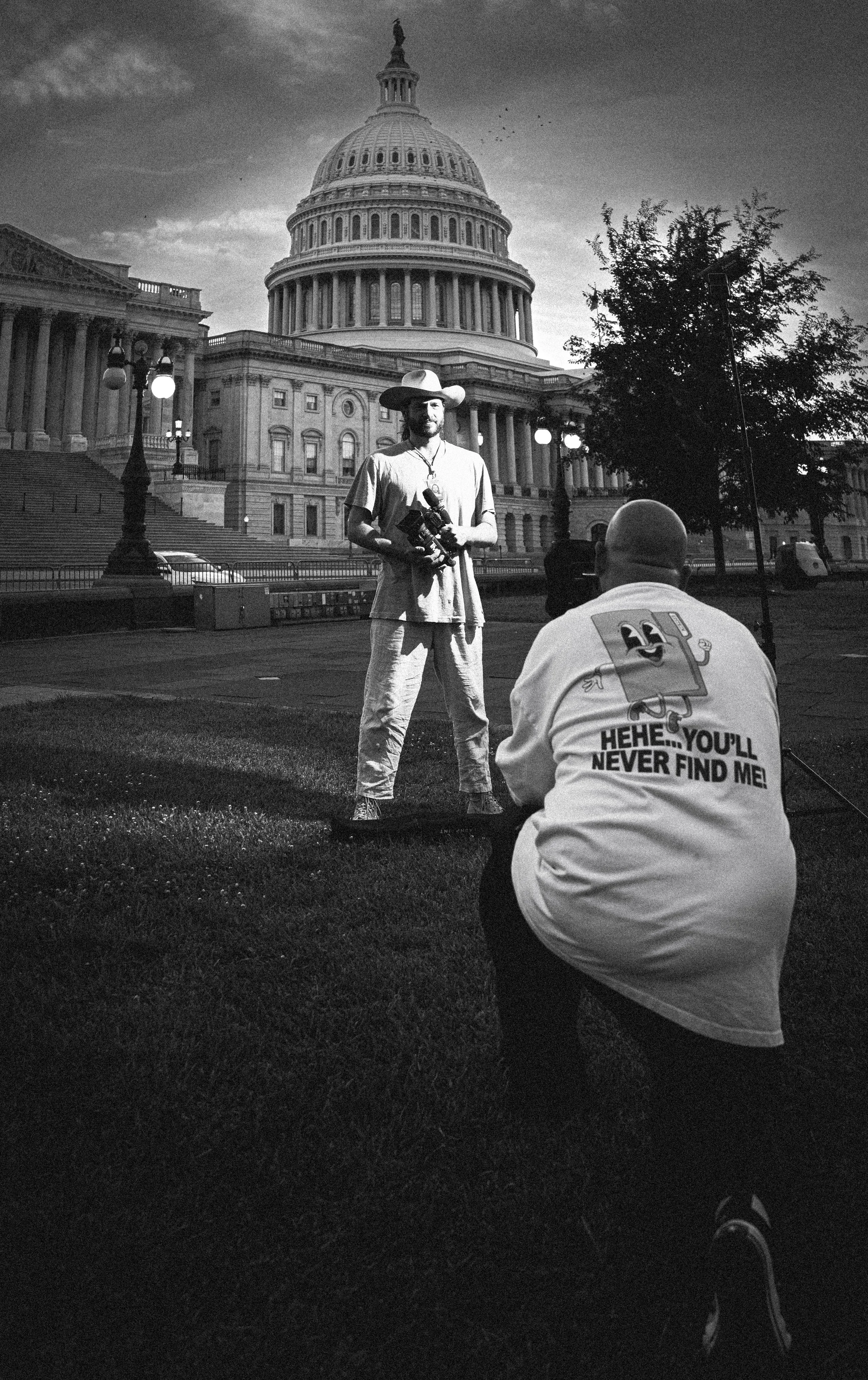 A man with a wide-brimmed hat and casual clothing stands outdoors holding a camera in front of the U.S. Capitol building at sunset. Another person in a white t-shirt with a cartoon character and the text "Hehe... you'll never find me!" is seated on t