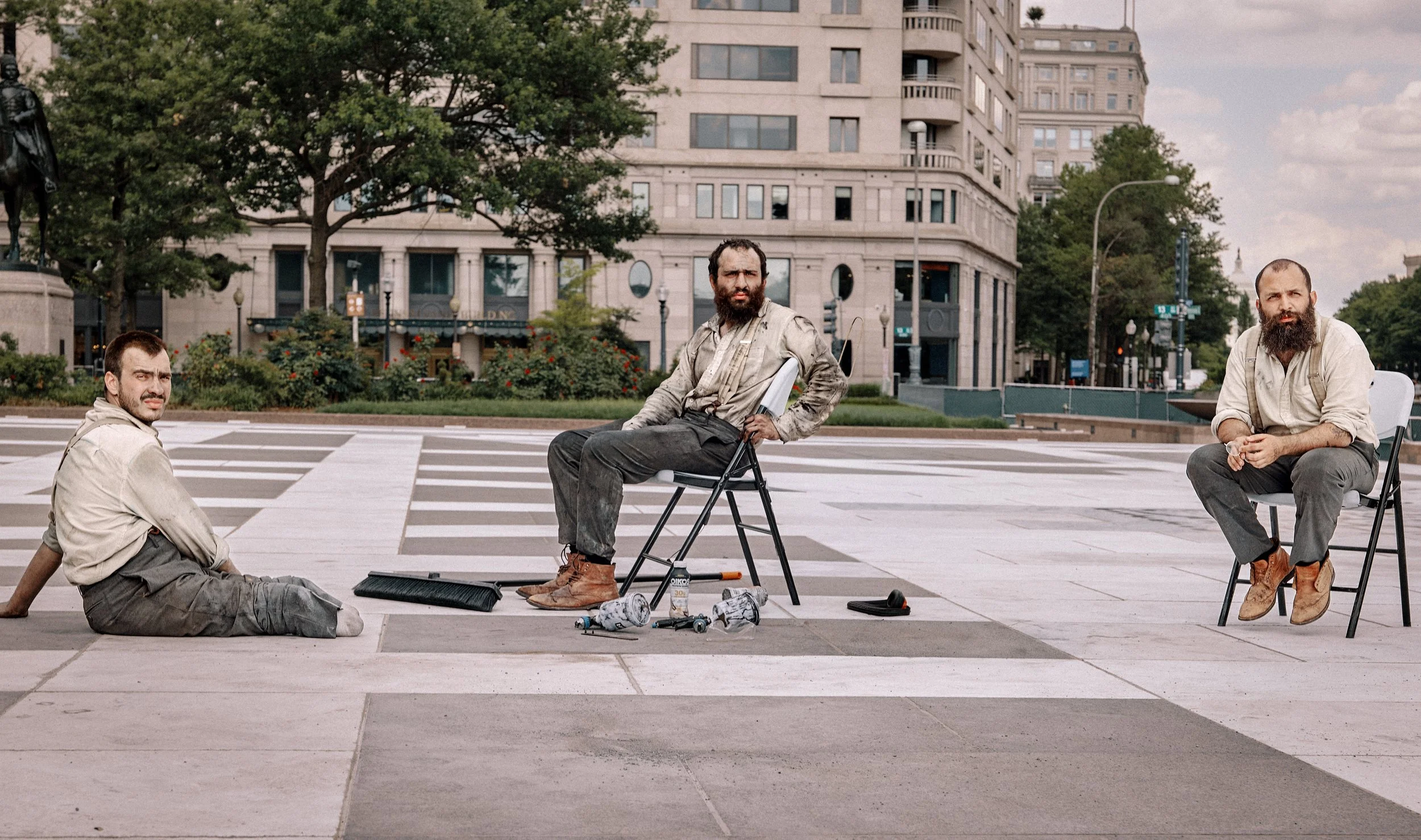 Three French sculptors dressed in dirty clothing sit on a city sidewalk with construction tools and debris scattered around them. They appear tired and are in an urban area with tall buildings and trees in the background. Freedom Plaza in DC.