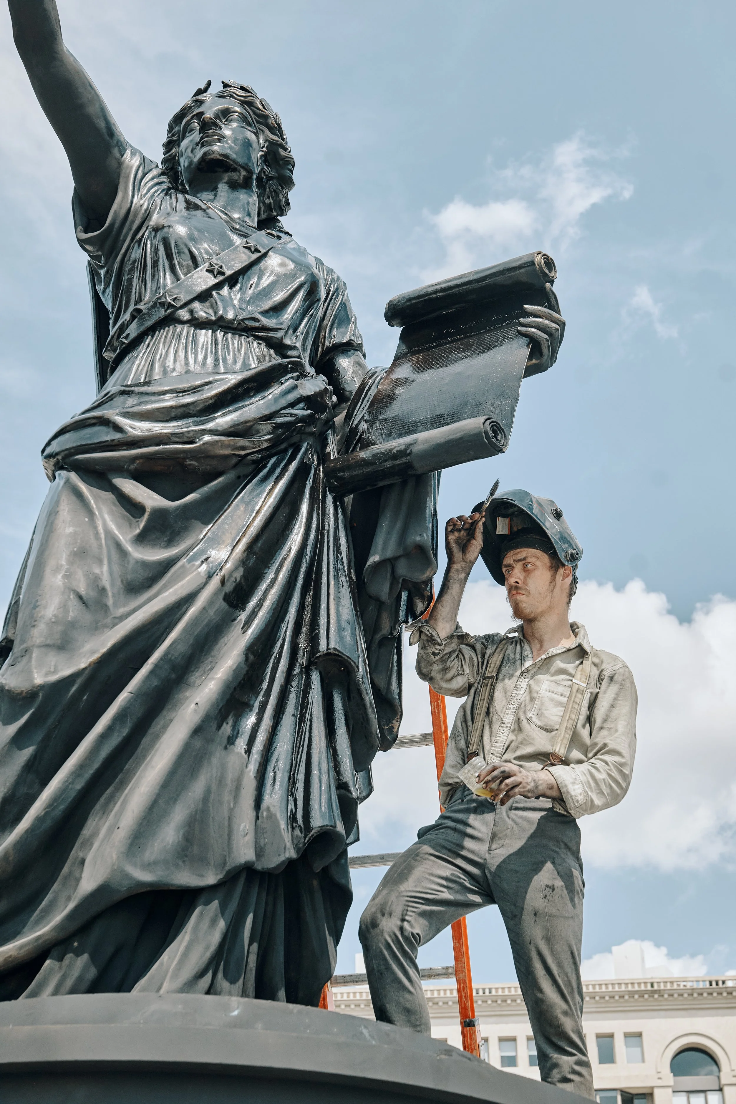 A worker in a beige uniform and helmet standing next to the Statue of Liberty, holding tools and working on it outdoors in a city setting.