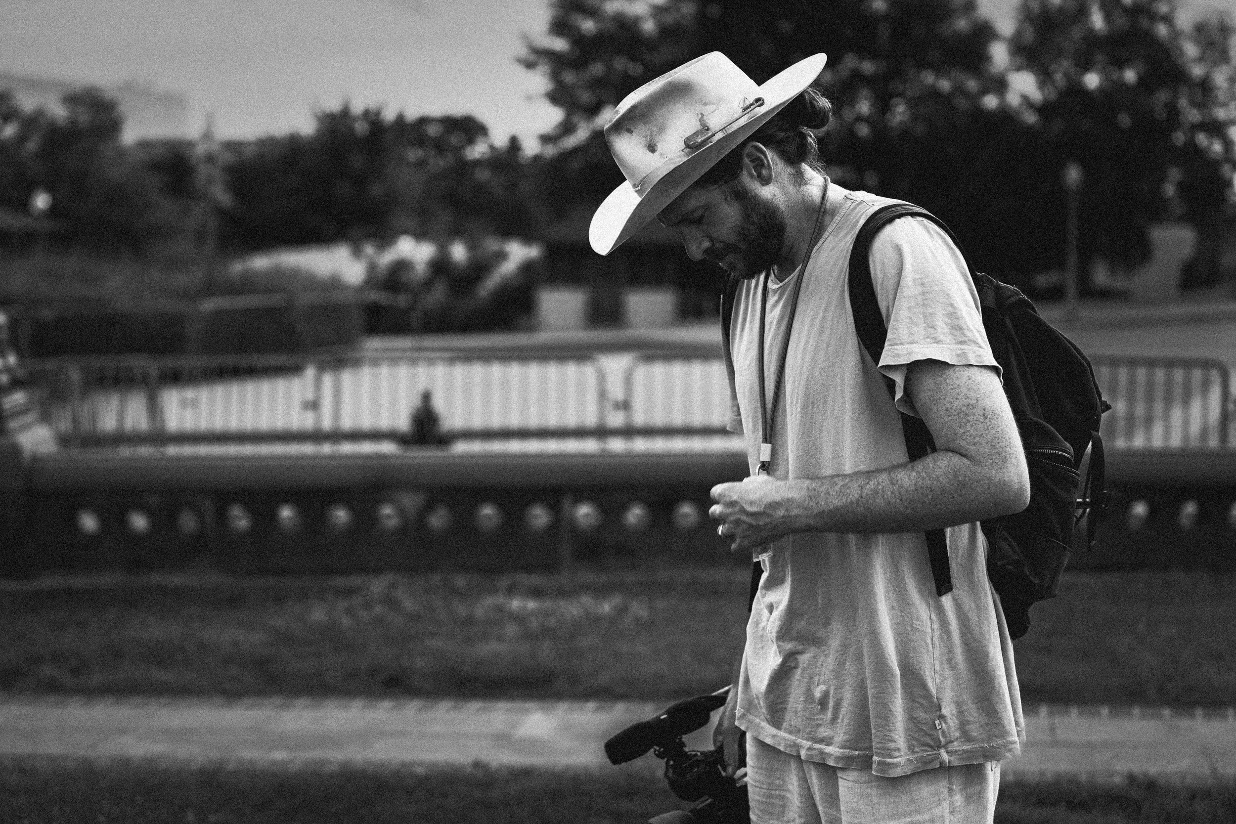 A man with a beard and long hair wearing a wide-brimmed hat and carrying a backpack, looking down at his phone while walking outdoors, with trees and a fence visible in the background.