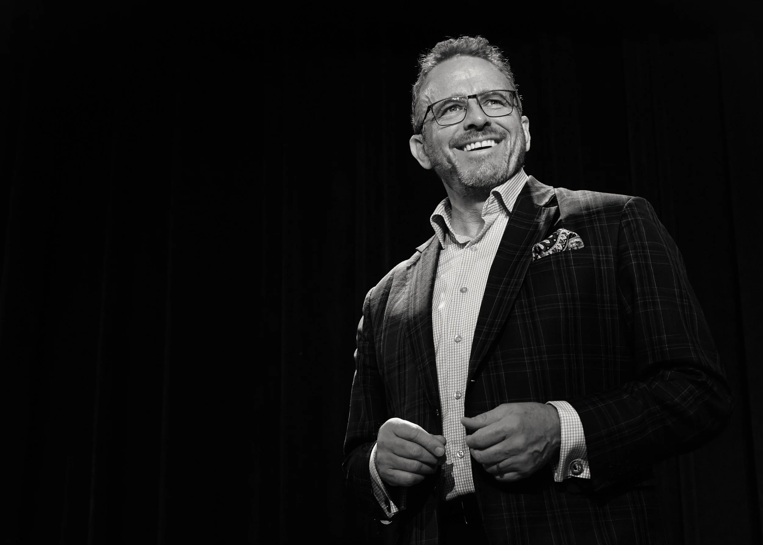 Black and white photo of a smiling man wearing glasses, a checked shirt, and a plaid blazer, standing against a dark background.