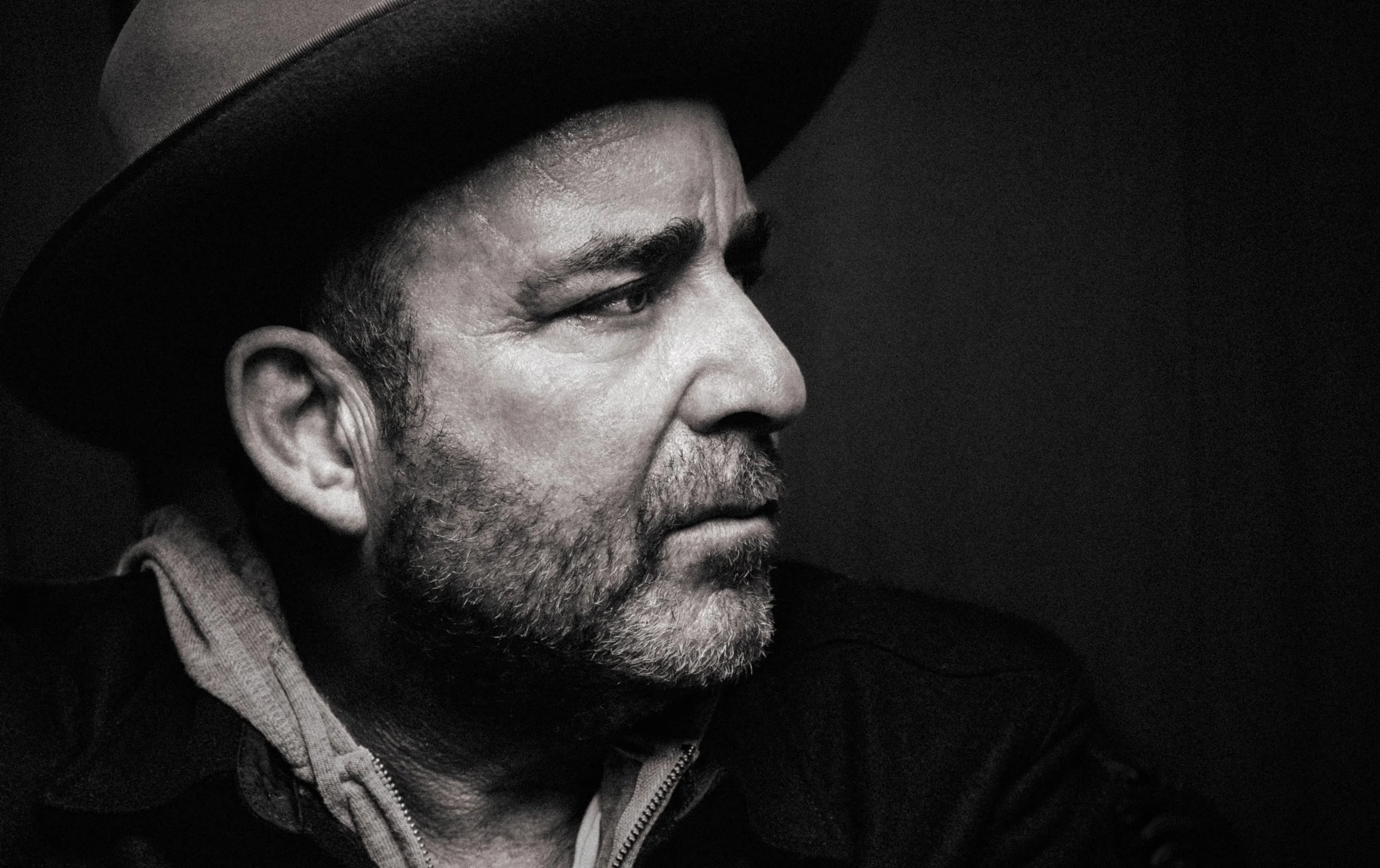 Profile portrait of a man with a beard, Jon Kahn, wearing a large hat and a jacket, against a dark background.
