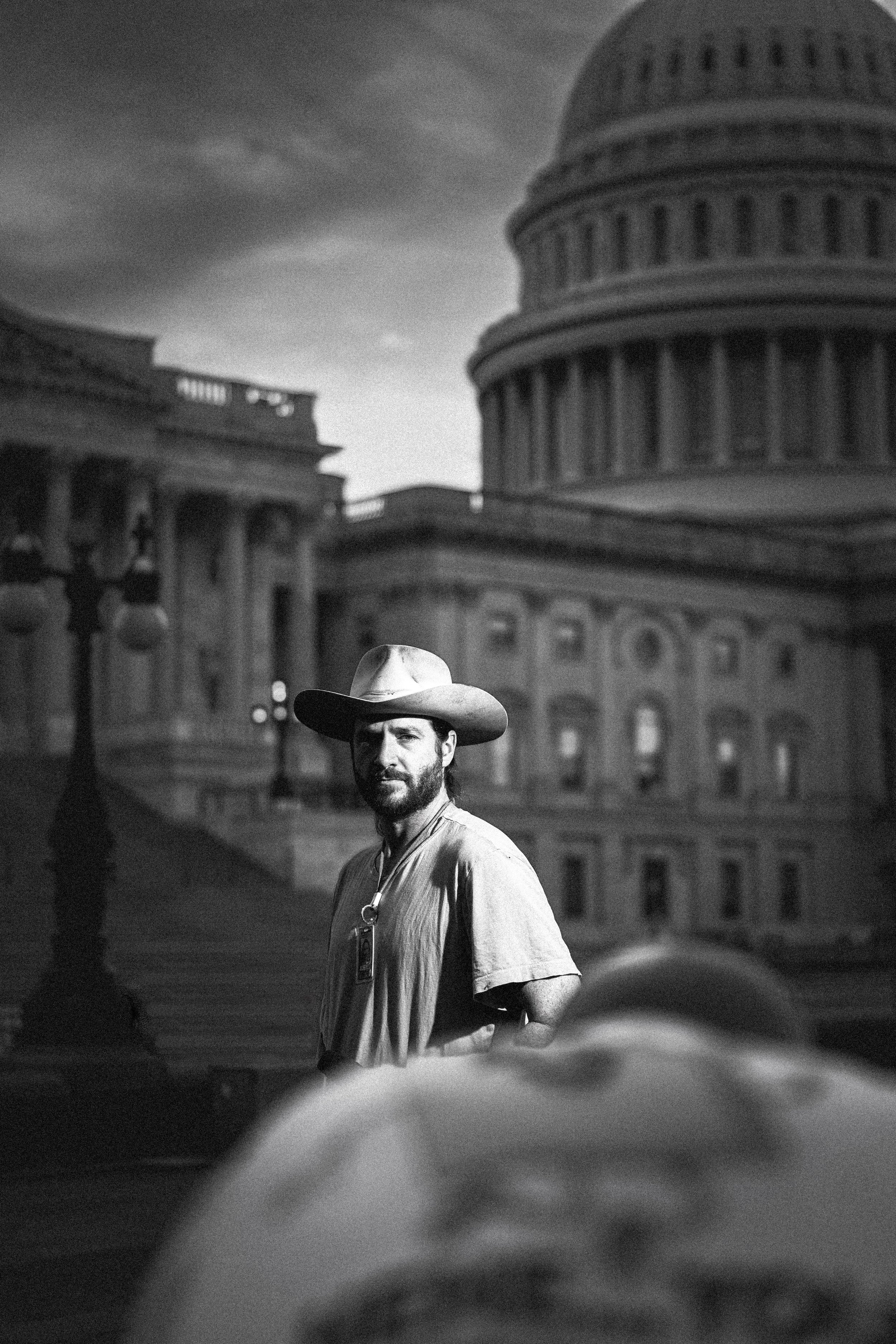 A man with a beard wearing a wide-brimmed hat and a badge around his neck stands in front of the U.S. Capitol building. The image is in black and white and has a blurry background with clouds.
