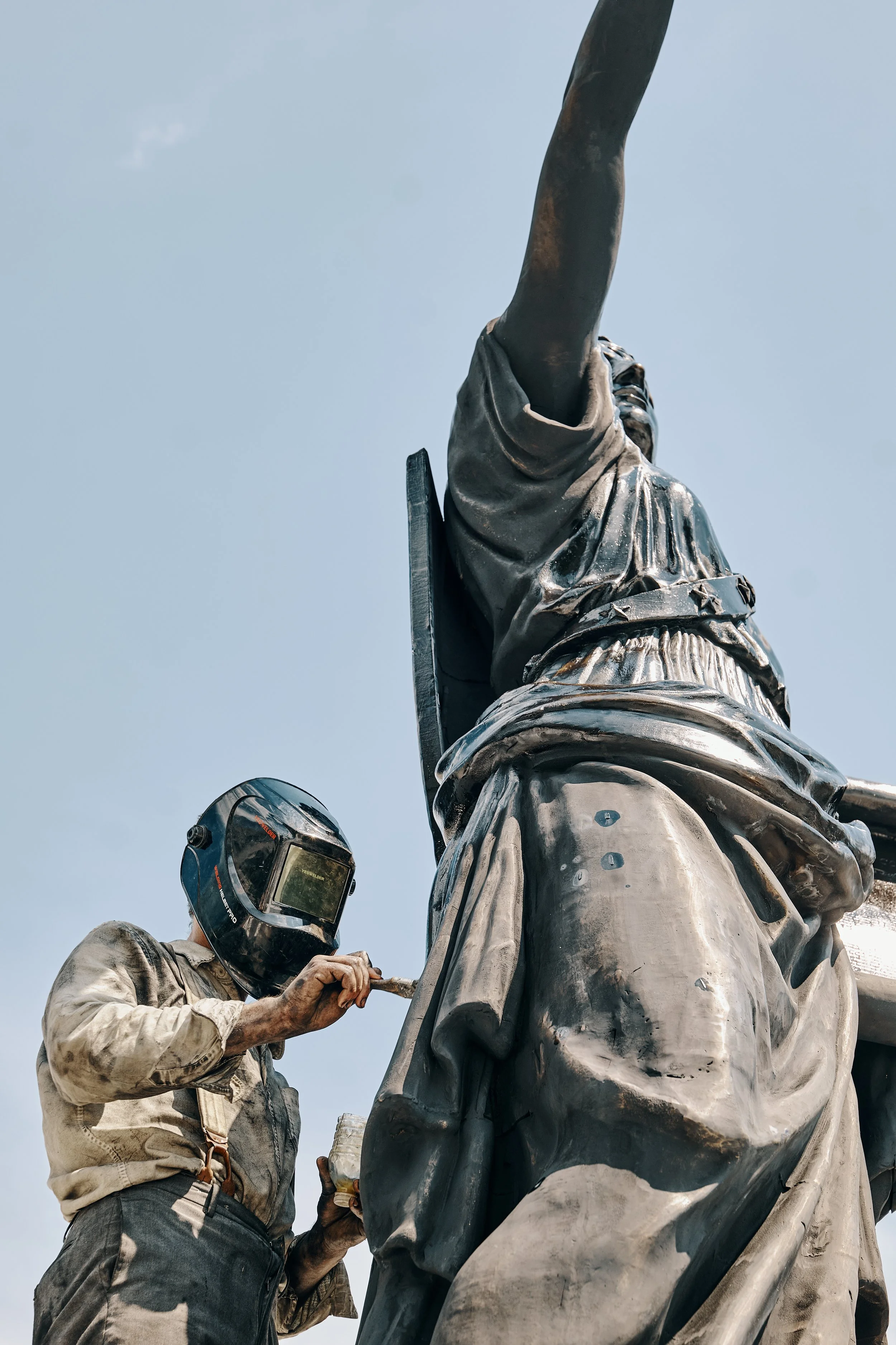 A French sculptor in protective gear installing a large bronze statue of a woman reaching upward against a clear blue sky.