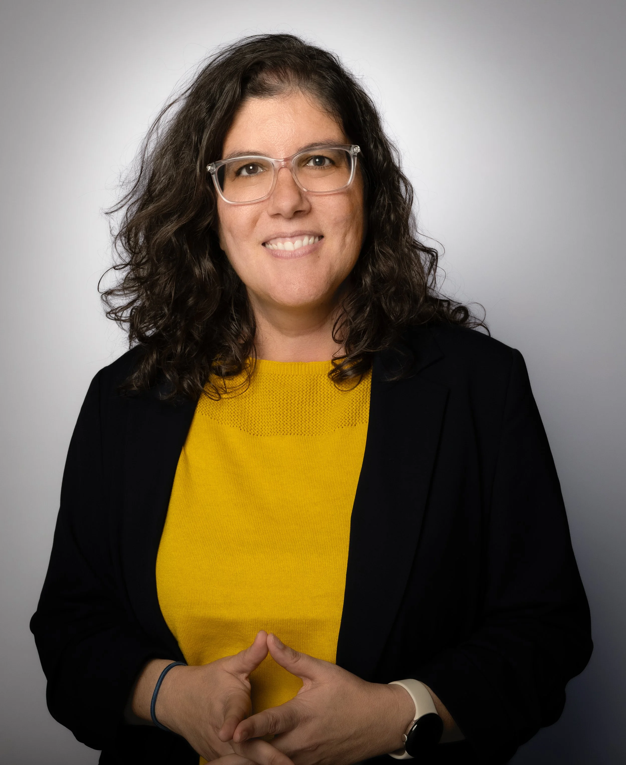 A woman with dark, curly hair wearing glasses, a yellow top, and a black blazer making a cautious hand gesture with intertwined fingers, standing against a plain gray background.