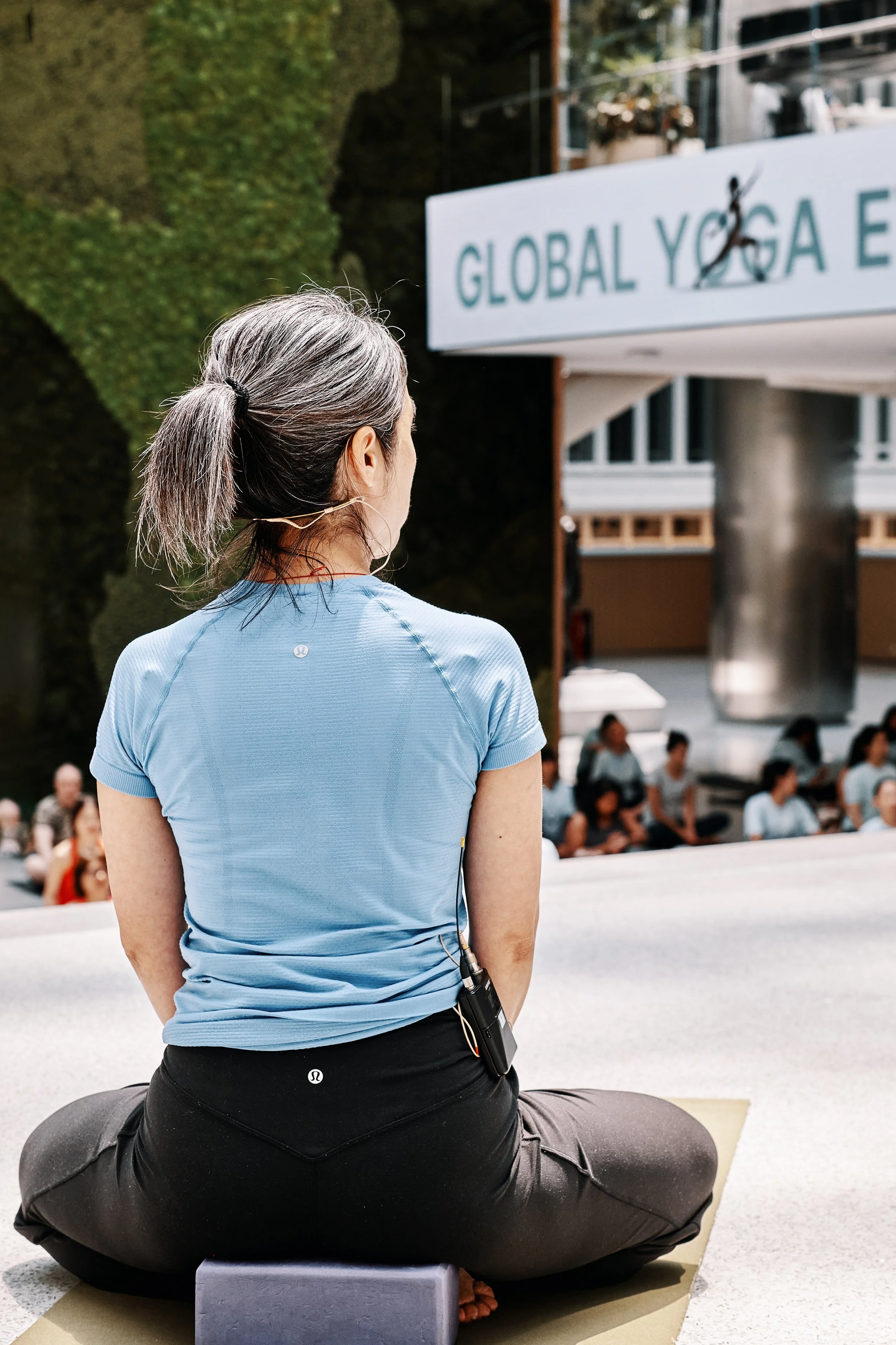 A woman with gray hair tied back, practicing yoga outdoors during daytime. She is seated cross-legged on a yoga mat, facing a large sign that reads 'GLOBAL YOGA'. In the background, there are other people sitting and the scene is surrounded by trees 
